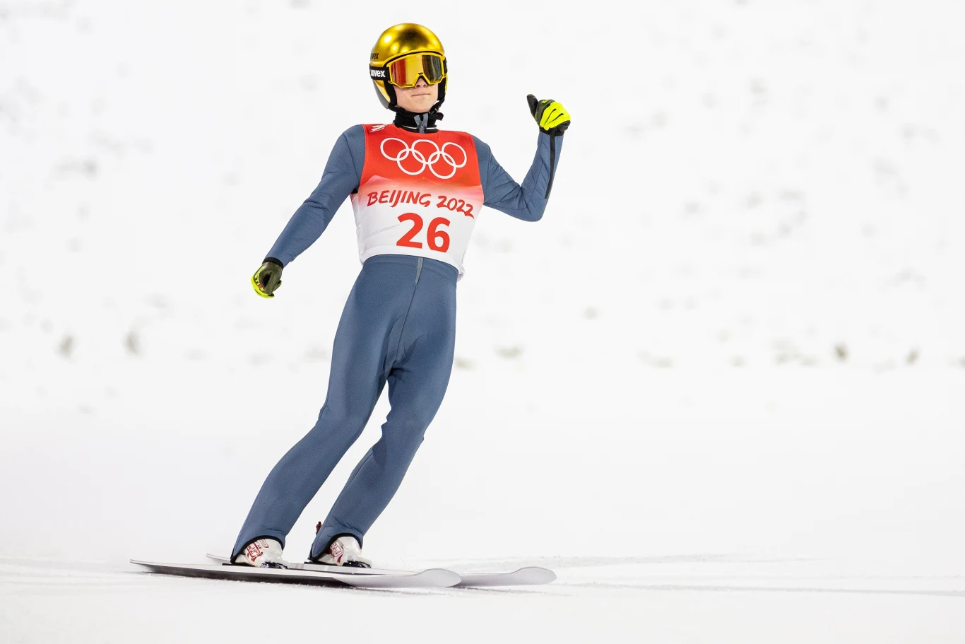 Skoczek narciarski w kombinezonie sportowym z numerem 26 ląduje na śniegu podczas Zimowych Igrzysk Olimpijskich Beijing 2022. Na głowie kask, na torsie plakietka olimpijska, w tle rozmyte tło pokryte śniegiem.