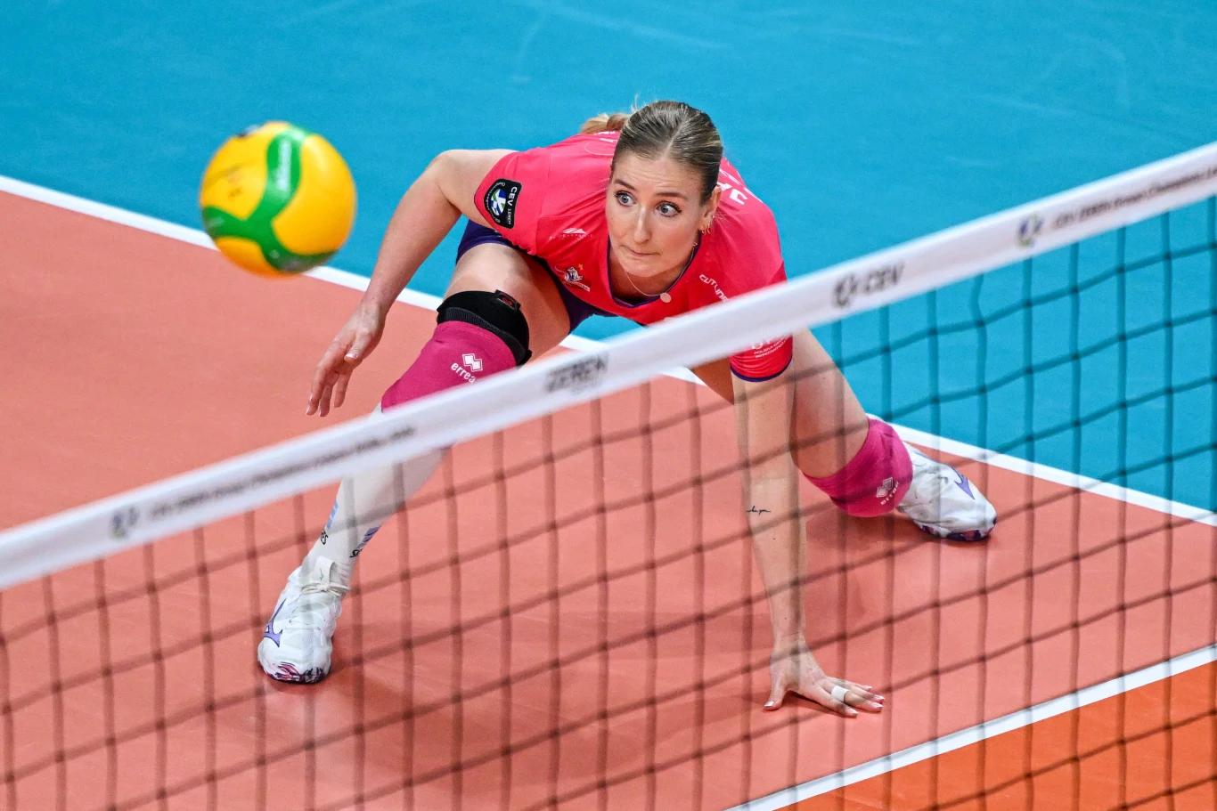 Siatkarka w różowym stroju wykonuje niską obronę podczas meczu, skupiona na nadlatującej piłce po drugiej stronie siatki, dynamiczna akcja na hali sportowej.