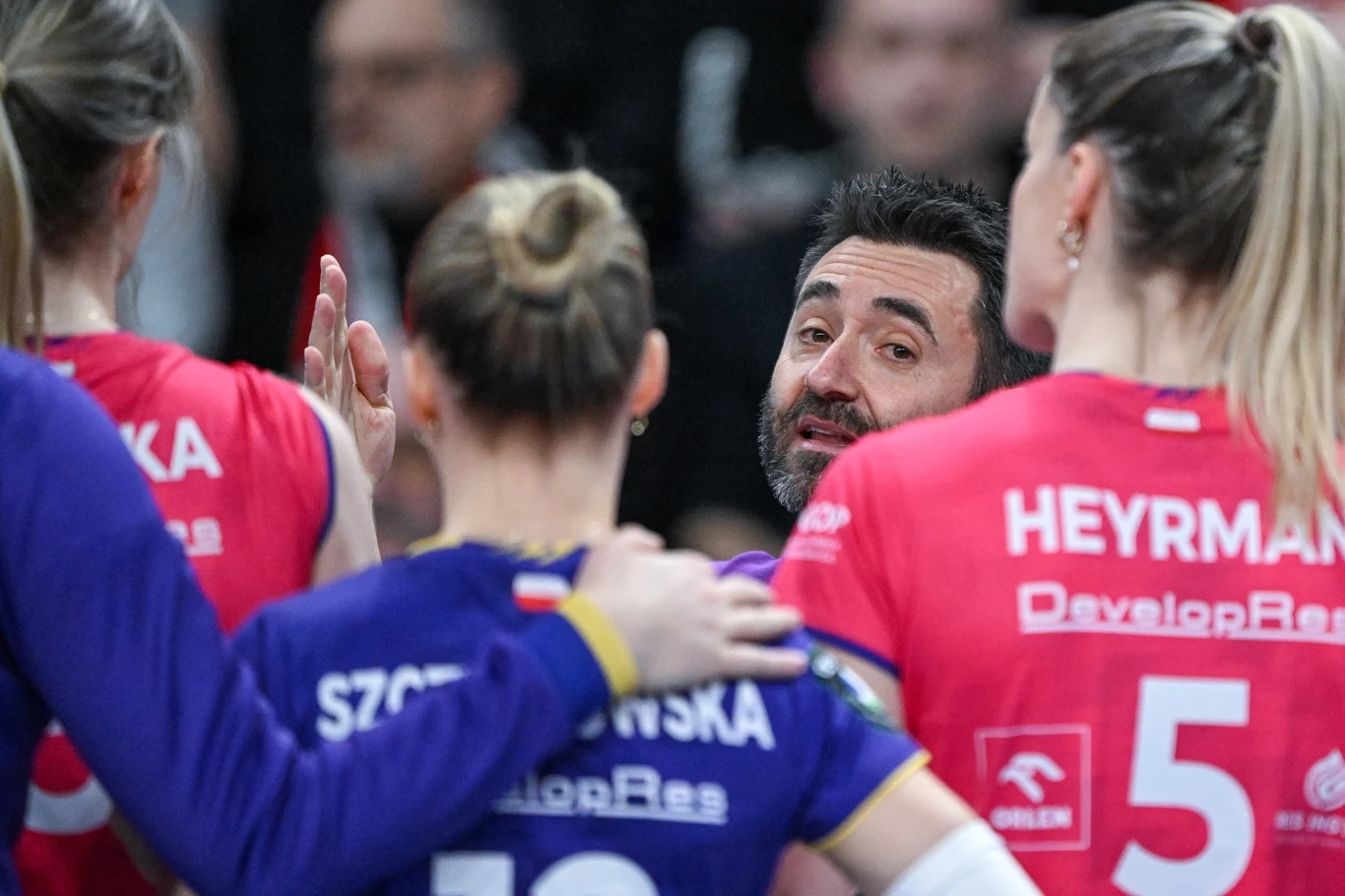 Grupa siatkarek w kolorowych strojach rozmawia z trenerem podczas meczu, w centrum uwagi znajduje się trener, który przekazuje wskazówki zawodniczkom, widoczne są nazwiska zawodniczek na plecach koszulek.