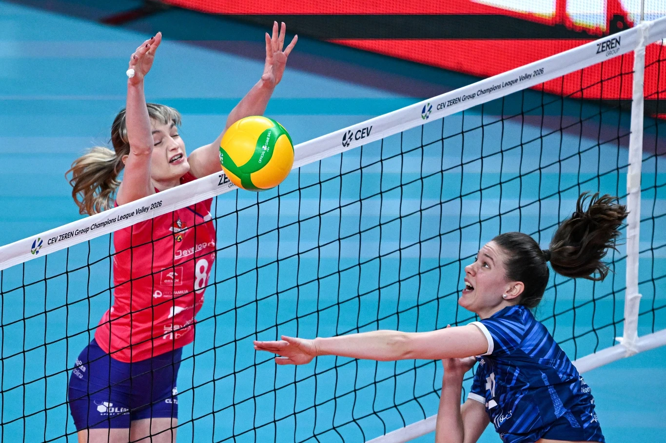 Dwie siatkarki podczas dynamicznego zagrania przy siatce, jedna w niebieskiej koszulce odbija piłkę w kierunku zawodniczki w czerwonym stroju wykonującej blok. Na tle widoczna siatka z oznaczeniami turnieju.