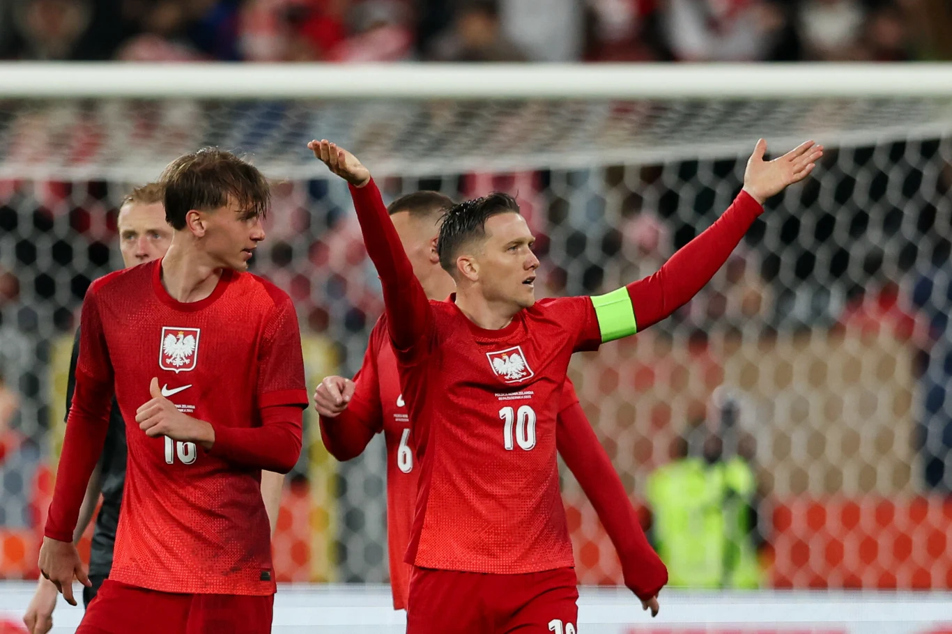 Dwóch piłkarzy reprezentacji Polski w czerwonych strojach podczas meczu na stadionie, jeden z nich z opaską kapitana ma uniesione ręce w geście niezadowolenia lub dyskusji.