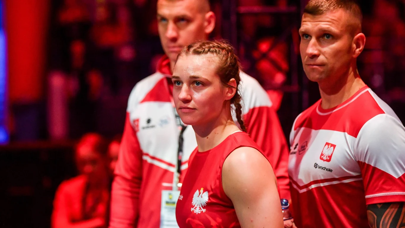 Kobieta ubrana w strój sportowy w barwach Polski stoi w otoczeniu dwóch mężczyzn w koszulkach z godłem Polski, atmosfera wskazuje na zawody sportowe lub wydarzenie reprezentacyjne.