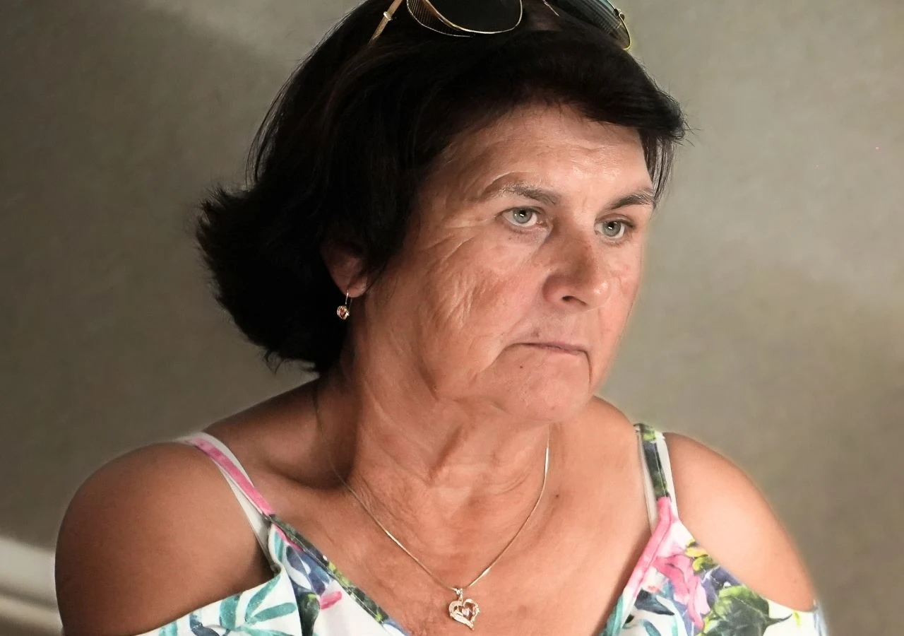 Grażyna Nowicka z "Sanatorium miłości" Starsza kobieta o ciemnych włosach i poważnym wyrazie twarzy, ubrana w kolorową bluzkę z odkrytymi ramionami i złoty naszyjnik z zawieszką w kształcie serca, na głowie ma okulary przeciwsłoneczne.