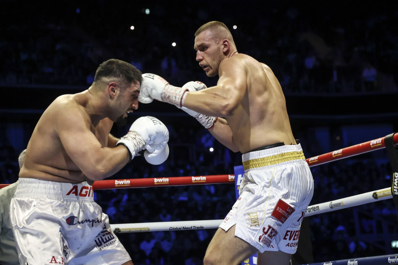 Dwóch bokserów w ringu podczas walki, ubrani w białe spodenki i rękawice, jeden z nich unika ciosu, drugi zadaje mocny prawy prosty, widownia i oświetlenie w tle podkreślają atmosferę zawodów.