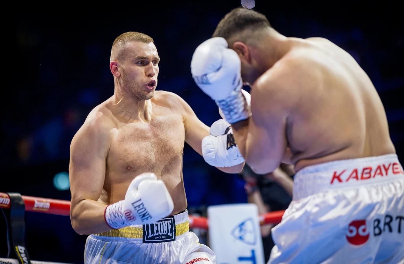 Dwóch bokserów w trakcie zaciętej walki na ringu, jeden z zawodników unosi ręce w obronnej postawie, drugi przygotowuje się do zadania ciosu, wokół nich widoczne liny ringu i rozmyte tło widowni.