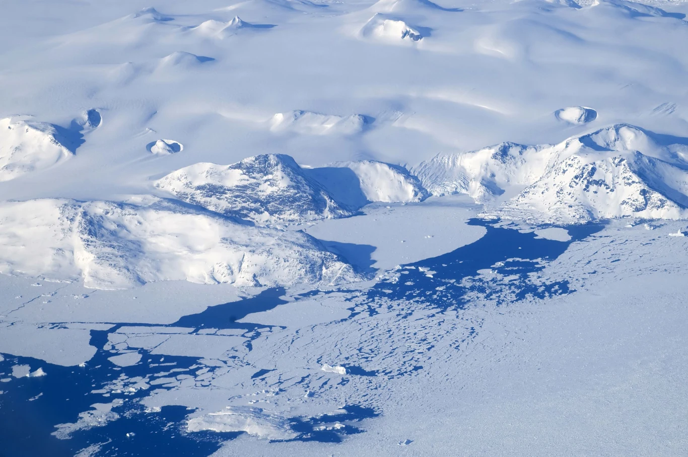 Rozległa pokrywa lodowa, fragmenty pękniętych tafli lodu oraz góry częściowo pokryte śniegiem i lodem. Widoczne kontrasty między białą powierzchnią lodu a ciemnoniebieską wodą w szczelinach.
