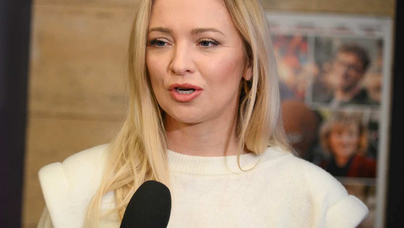 Barbara Kurdej-Szatan Kobieta o blond włosach ubrana w białą bluzę, wypowiada się do mikrofonu. W tle widoczny rozmazany plakat z postaciami.