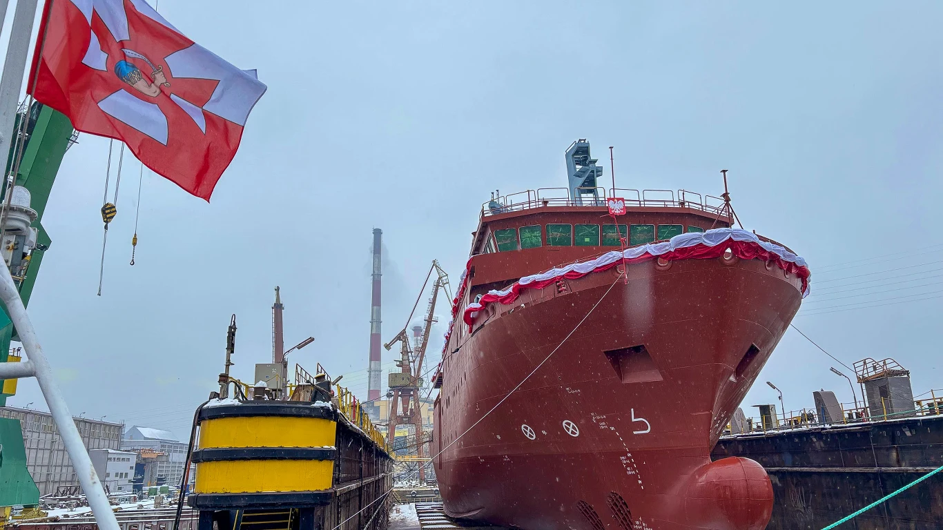 Nowo wybudowany, duży statek zacumowany w doku stoczniowym na tle żurawi i przemysłowych instalacji, z przodu widoczna czerwona bandera z białym symbolem.