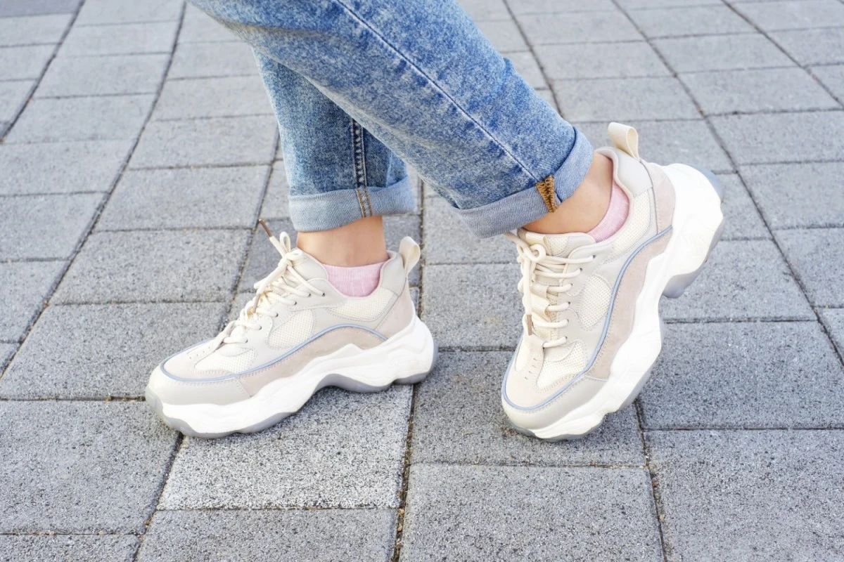 New Balance – poznaj kultowe modele, które warto mieć w szafie Białe sportowe buty na grubej podeszwie, nogi w jasnych jeansach, szara kostka brukowa.