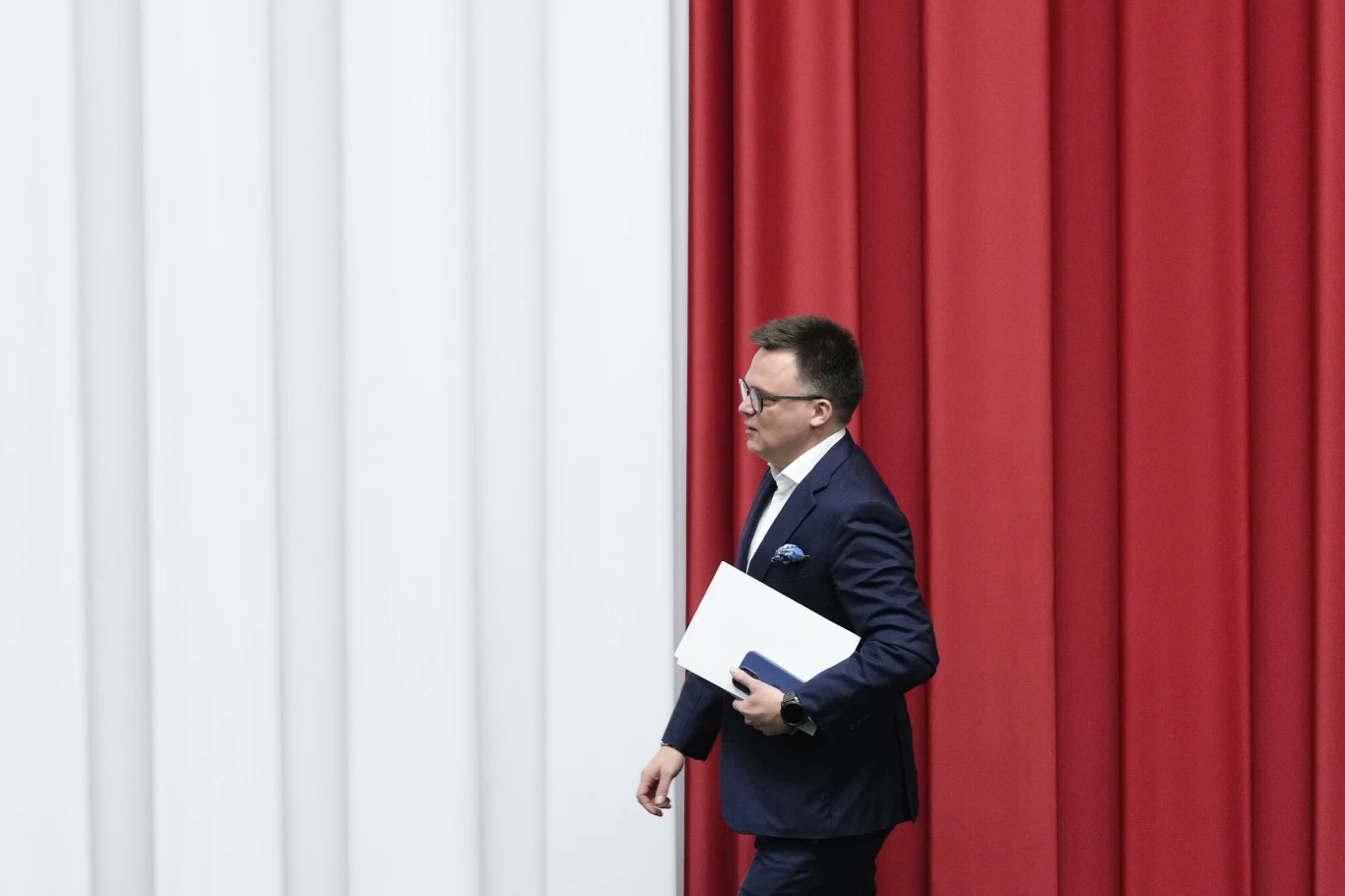 Szymon Hołownia niespodziewanie wróci do gry? Mężczyzna w garniturze idzie obok dużej ściany w barwach narodowych Polski, trzymając dokumenty i tablet w rękach.
