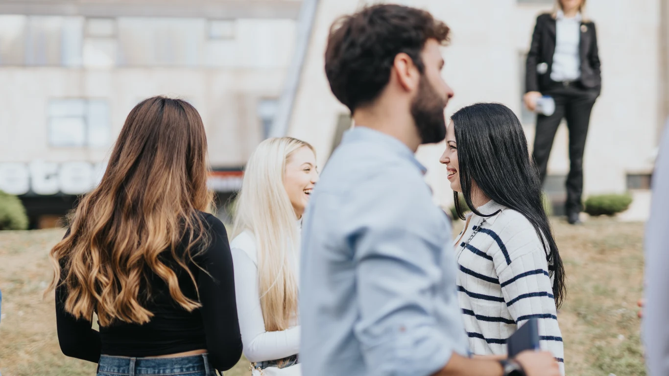 Młodzi dorośli wyróżniają się w statystykach dotyczących spożycia alkoholu (zdj. ilustracyjne) Grupa młodych osób rozmawia ze sobą na świeżym powietrzu, w tle widoczna kobieta w garniturze stojąca w oddali.
