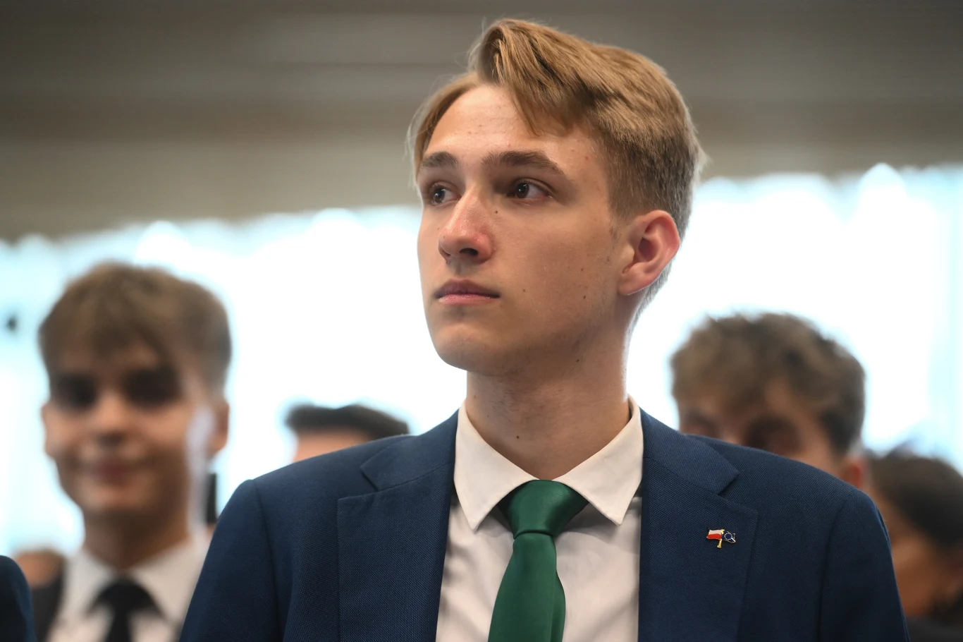 Paweł Mrozek, Akcja Uczniowska Młody mężczyzna w eleganckim garniturze z zielonym krawatem, spoglądający poważnie przed siebie, w tle kilku innych młodych mężczyzn o rozmytych twarzach.