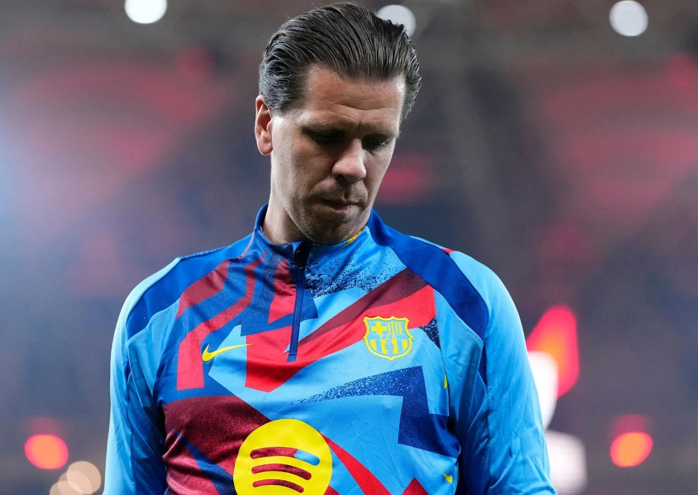 Wojciech Szczęsny Piłkarz w kolorowym stroju sportowym FC Barcelony patrzący w dół, światła stadionu rozmyte w tle.