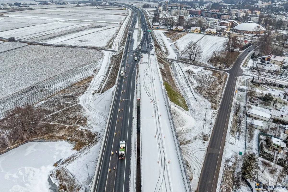 GDDKiA poinformowało o otwarciu nowego zjazdu na węźle Mistrzejowice. Zimowy krajobraz z drogą ekspresową przebiegającą przez ośnieżone pola, po której poruszają się samochody, po bokach widoczne są boczne drogi, zabudowania oraz fragment zamarzniętego zbiornika wodnego.