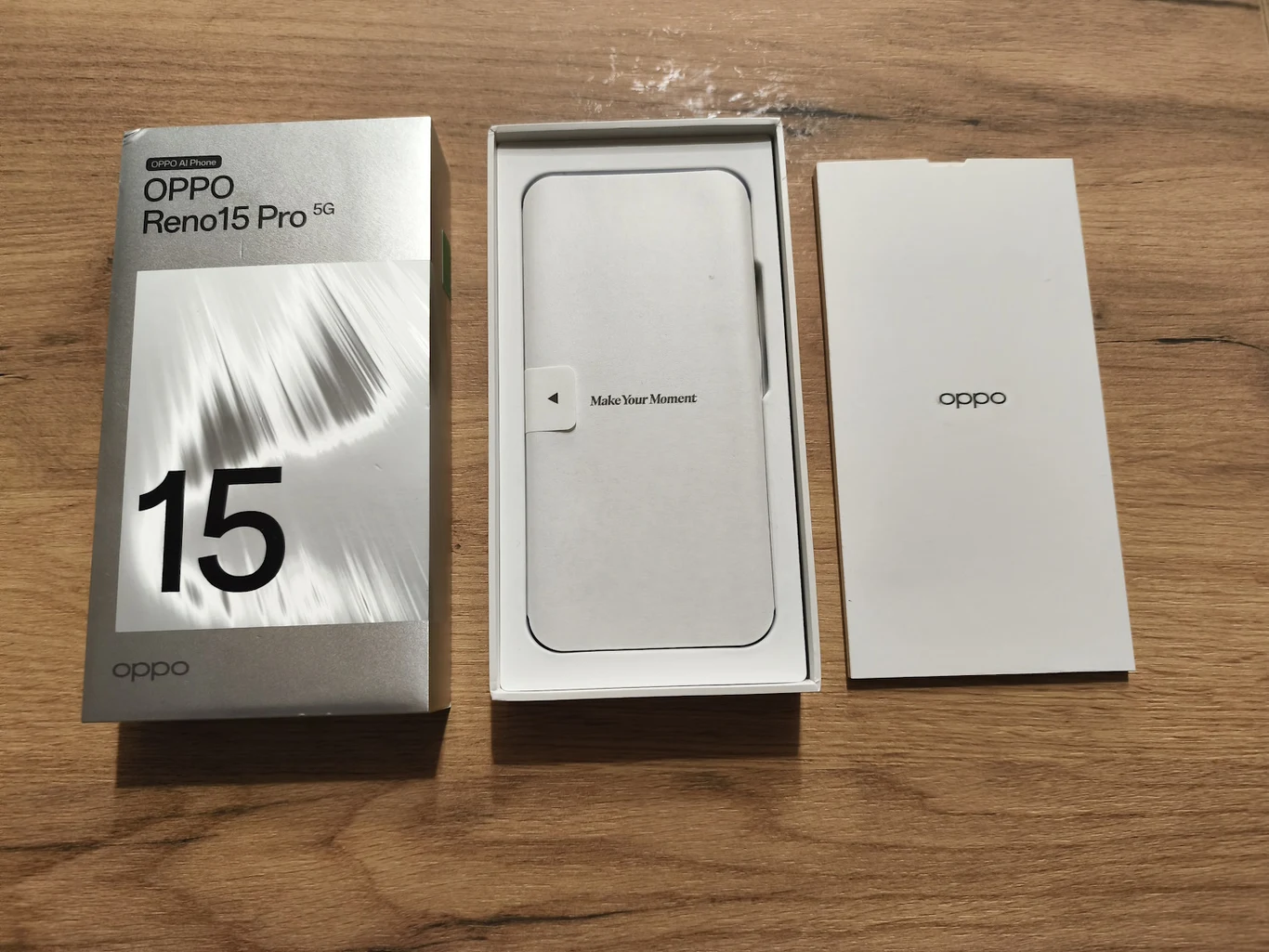 Oryginalne pudełko smartfona OPPO Reno15 Pro 5G leży na drewnianym blacie. Po prawej stronie pudełka znajduje się jego zawartość, w tym miejsce na telefon oraz papierowa wkładka z logo marki.
