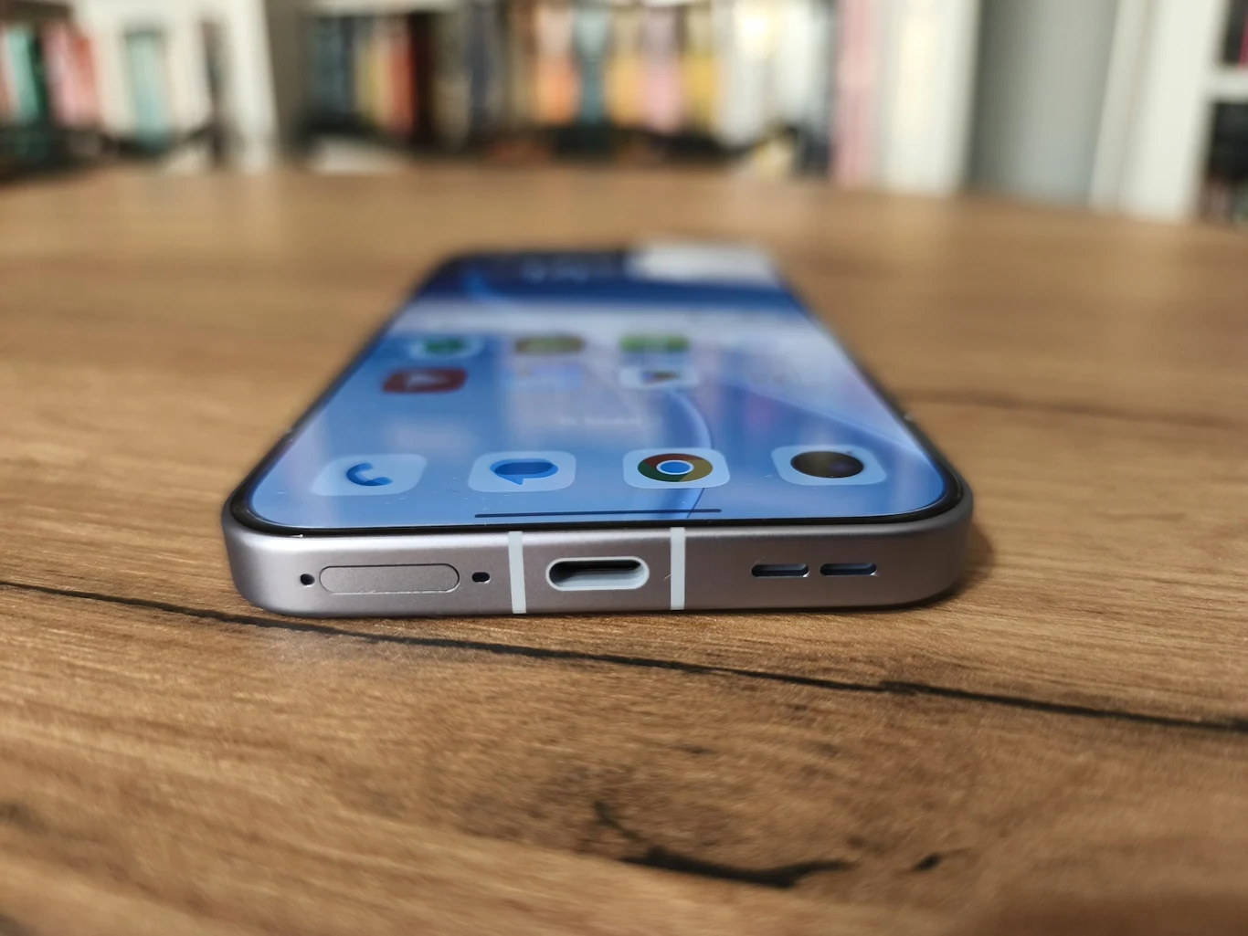 Nowoczesny smartfon leżący płasko na drewnianym blacie, ekran urządzenia wyraźnie wyświetla ikony aplikacji, a na pierwszym planie widoczny port ładowania USB-C oraz przyciski boczne obudowy.