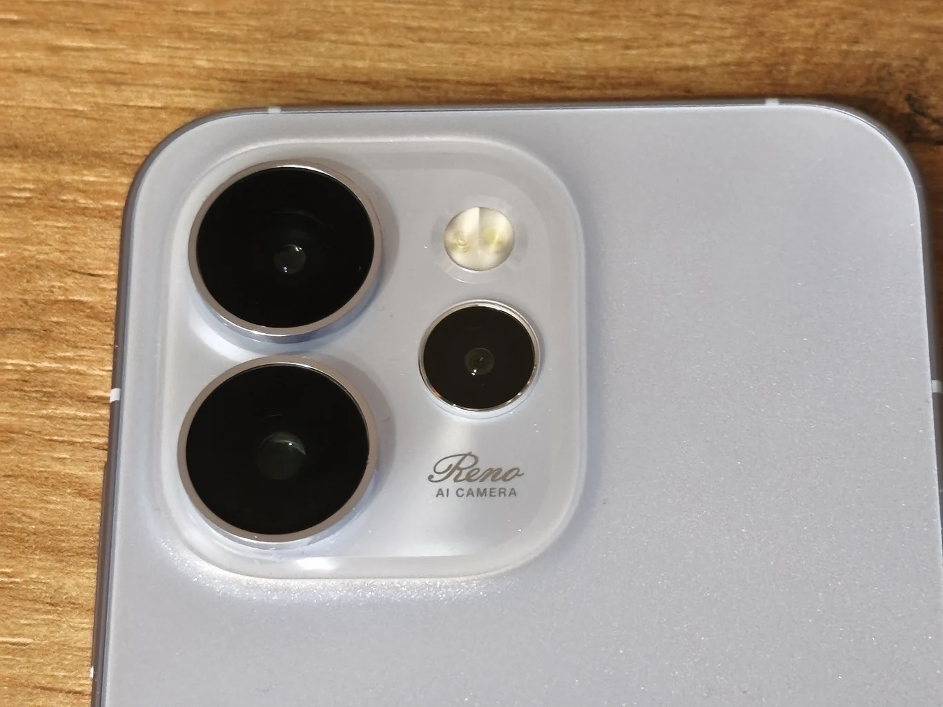 Zbliżenie na aparat tylni smartfona z trzema obiektywami oraz diodą doświetlającą na srebrnej obudowie. Widoczny napis Reno AI Camera. Tło drewniane.