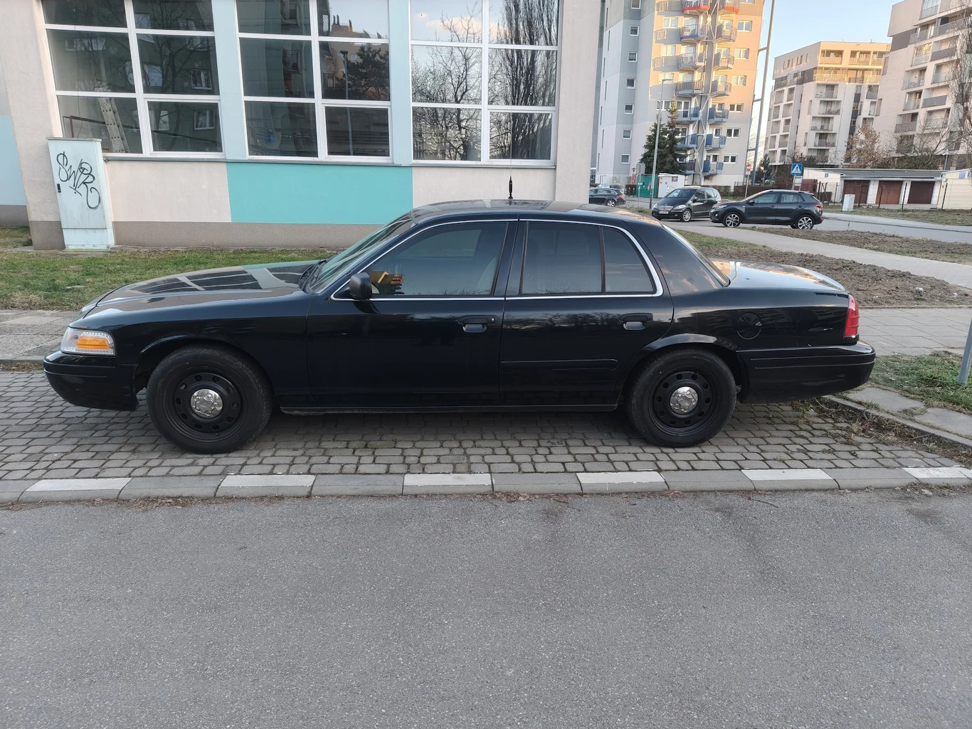 Czarny samochód osobowy typu sedan zaparkowany przy chodniku na tle budynku mieszkalnego i bloków. Szyby są mocno przyciemnione, a auto nie posiada widocznych oznaczeń sugerujących konkretnego właściciela lub instytucję.