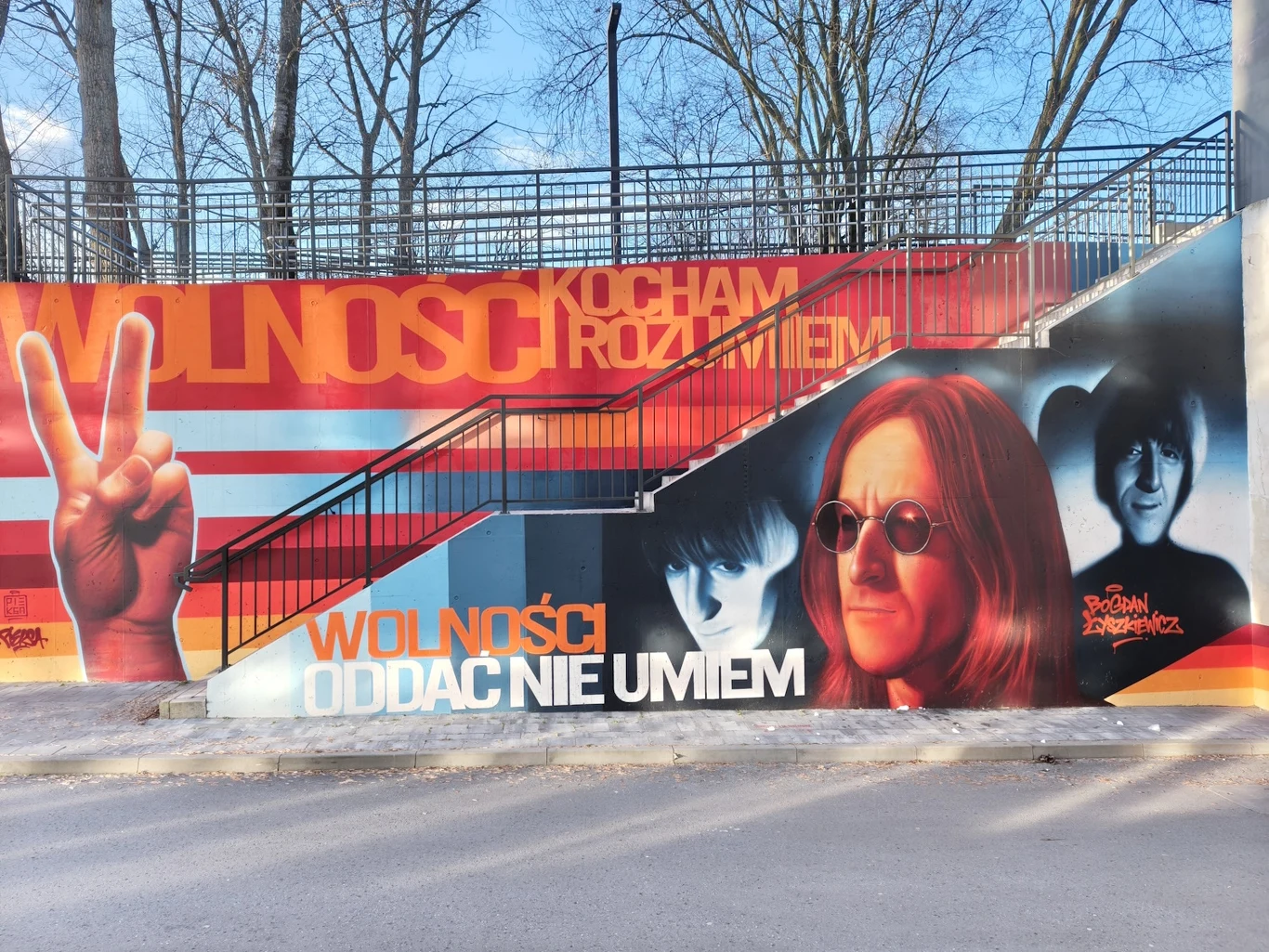 Kolorowy mural na ścianie betonowej przedstawia duże twarze osób, gest dłoni pokazującej znak pokoju oraz napisy o wolności i miłości. W tle widoczne są drzewa oraz stalowa balustrada schodów z czarną poręczą.
