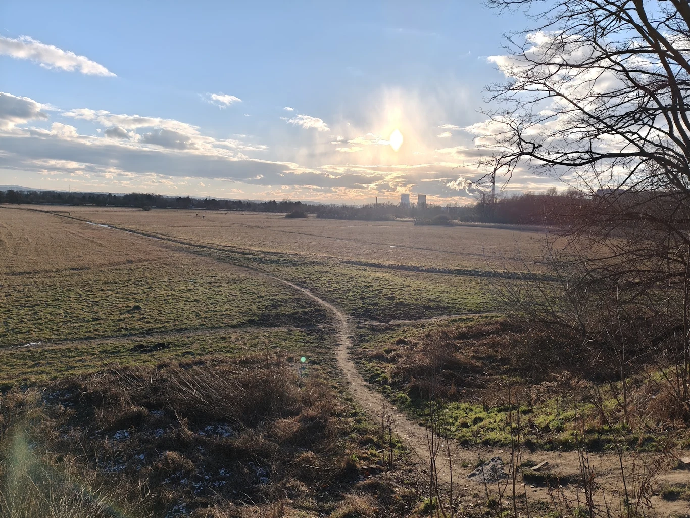 Polna ścieżka wijąca się przez rozległe, suche pole, oświetlona nisko świecącym słońcem, po prawej stronie widoczne bezlistne gałęzie drzewa, na horyzoncie widać zabudowania i kłęby pary lub dymu.