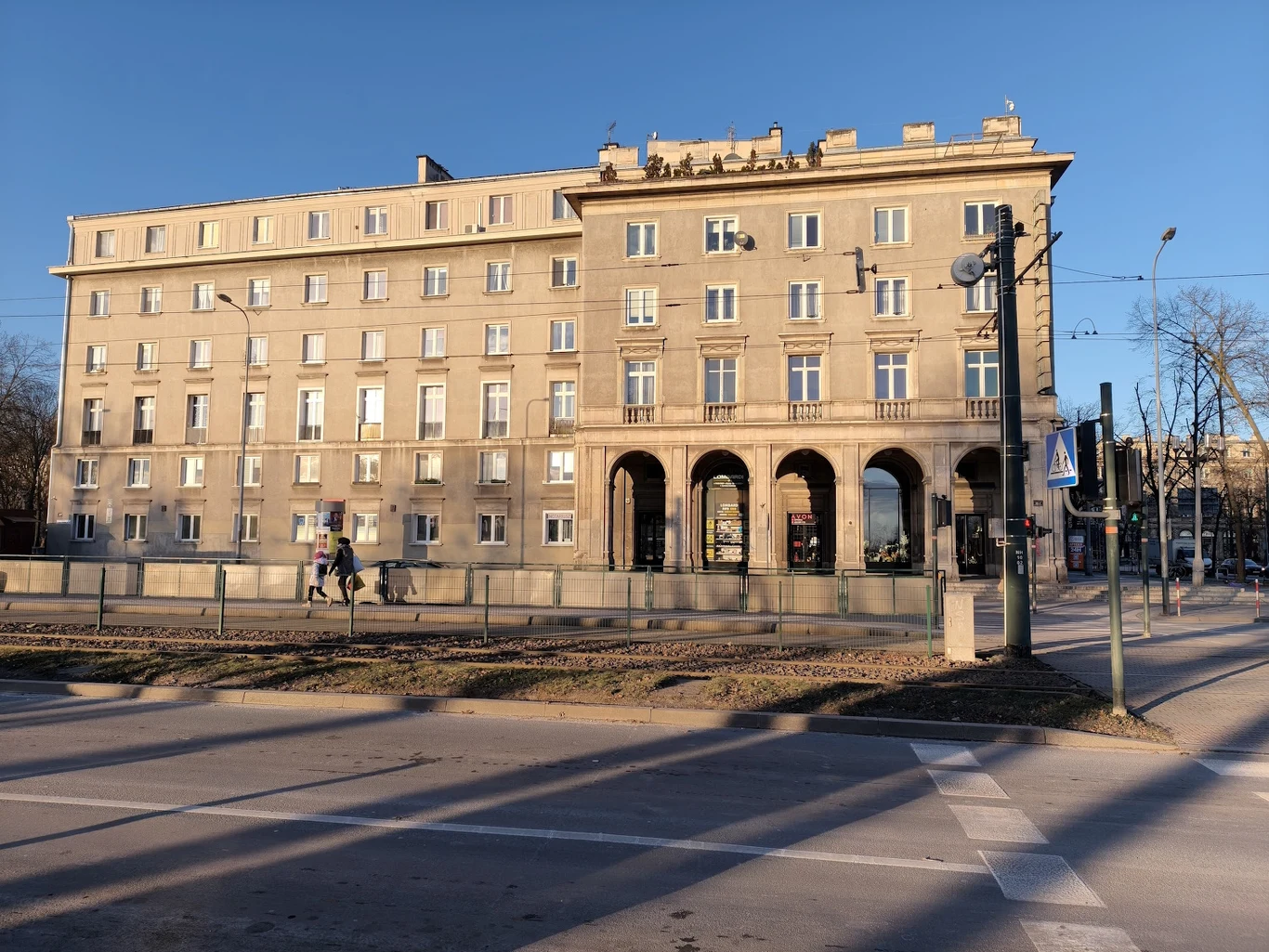 Duży, jasny budynek z licznymi oknami i arkadami na parterze zlokalizowany przy ruchliwej ulicy, po której spacerują ludzie, a latarnie rzucają długie cienie na jezdnię.