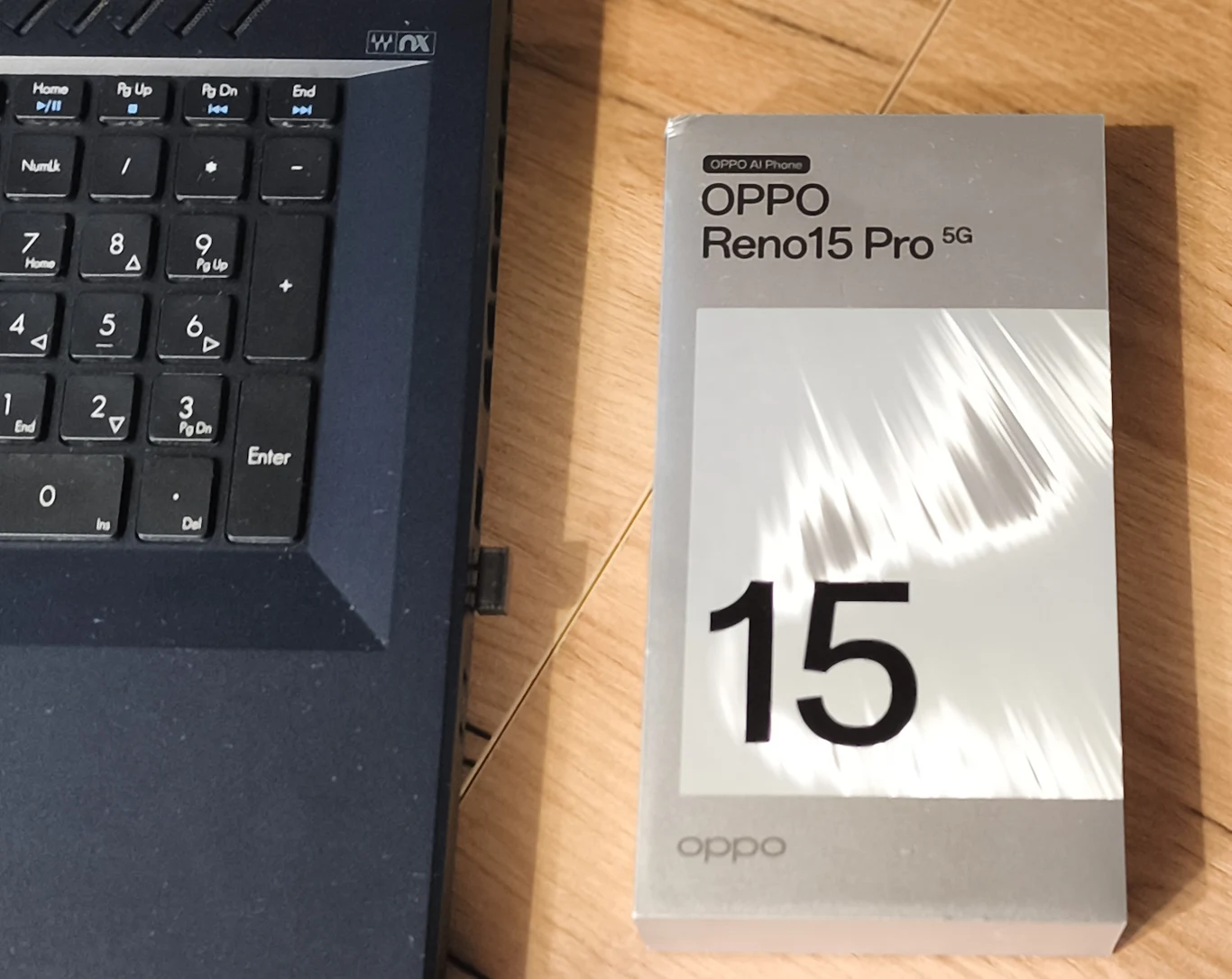 Nowoczesne pudełko smartfona OPPO Reno15 Pro 5G ustawione na drewnianej powierzchni obok klawiatury ciemnego laptopa, fragment laptopa ukazujący część klawiszy i touchpad.