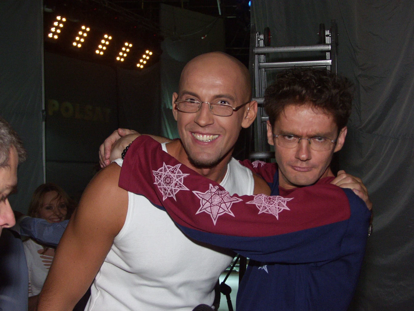 Dwóch mężczyzn pozuje razem, jeden obejmuje drugiego, obaj mają uśmiechy na twarzach, a w tle widoczne są elementy sceny i oświetlenie.