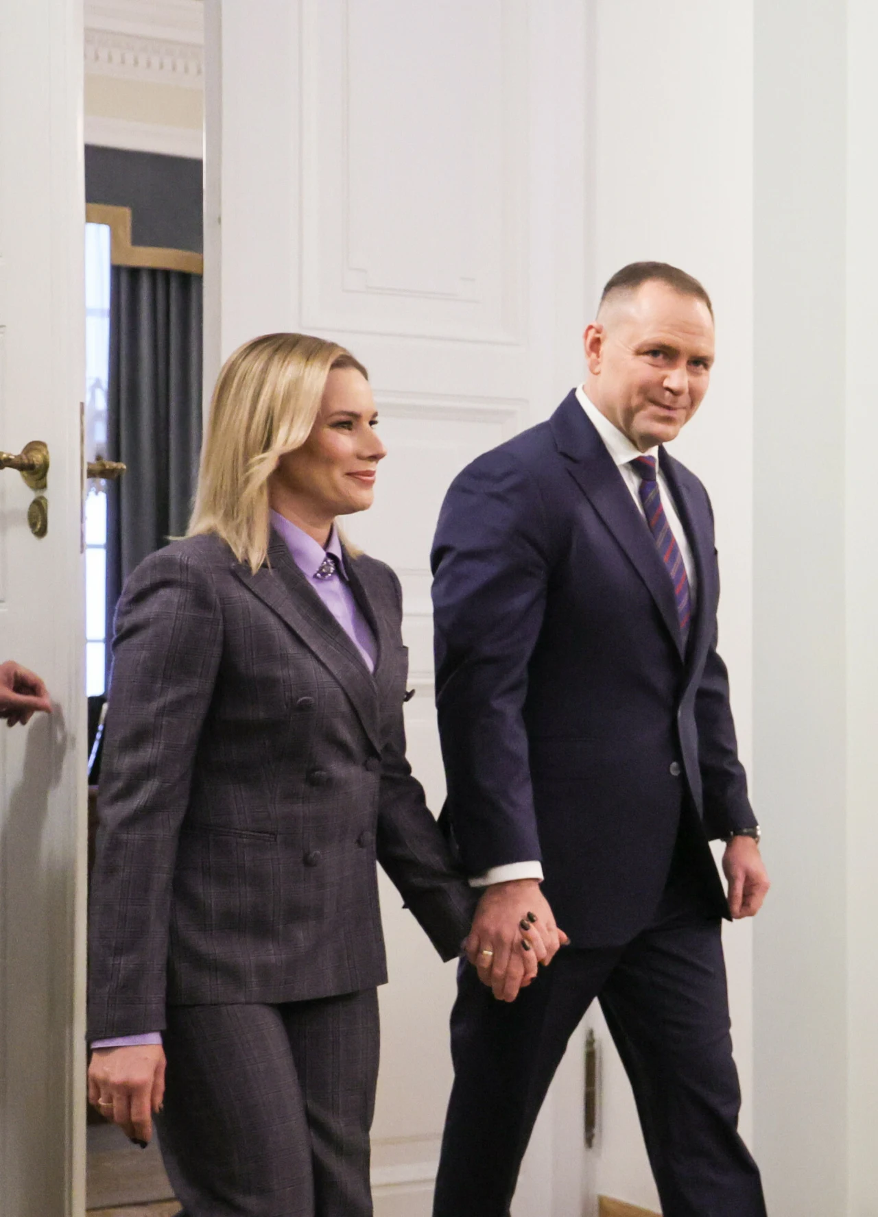 Elegancko ubrana kobieta i mężczyzna w garniturach wchodzą razem do jasnego pomieszczenia, trzymając się za ręce. Oboje wyglądają na pewnych siebie i uśmiechniętych.