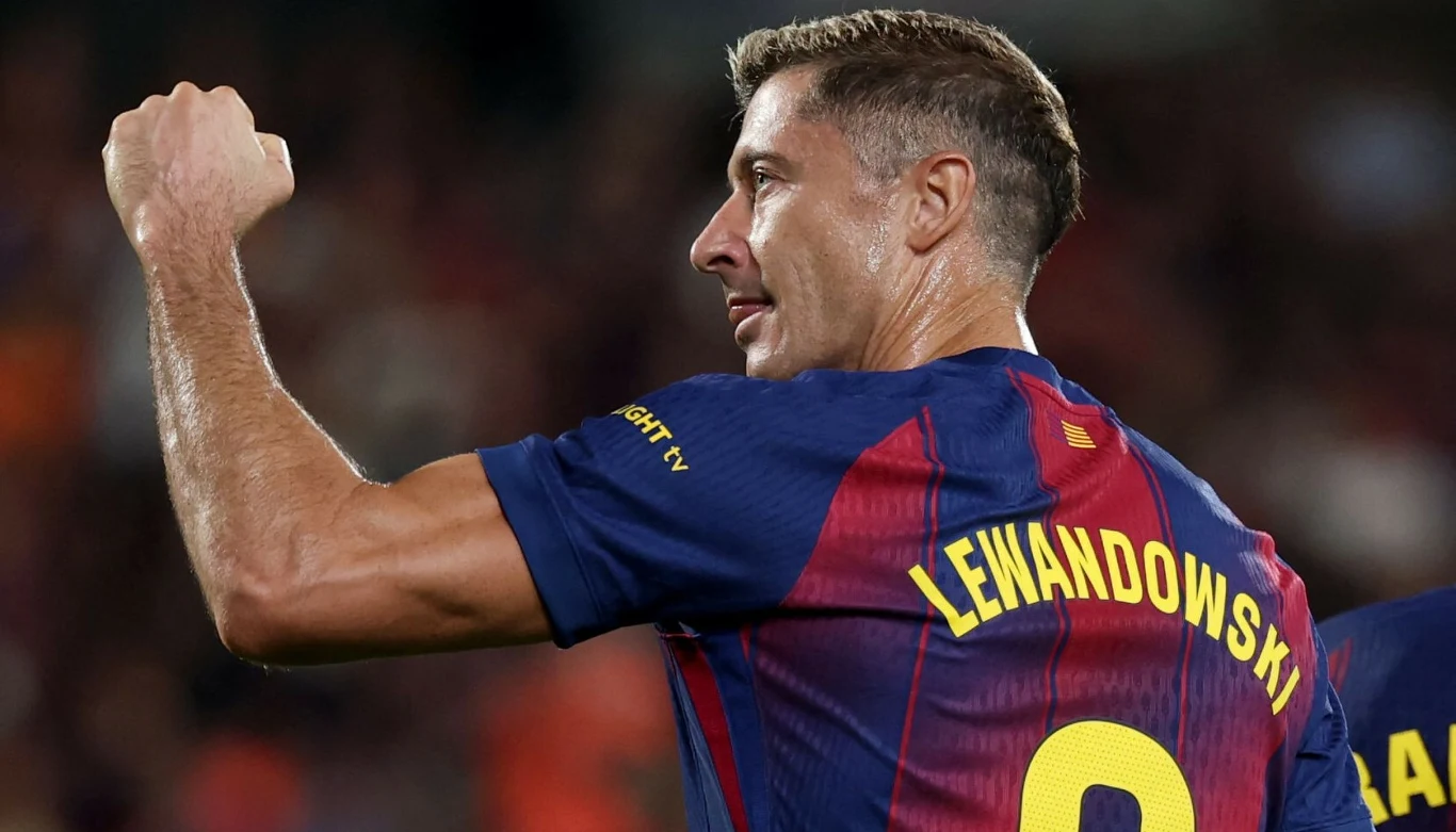 Piłkarz w granatowo-bordowej koszulce z nazwiskiem Lewandowski i numerem 9, unosi zaciśniętą pięść w geście triumfu na tle rozmazanej widowni stadionu.
