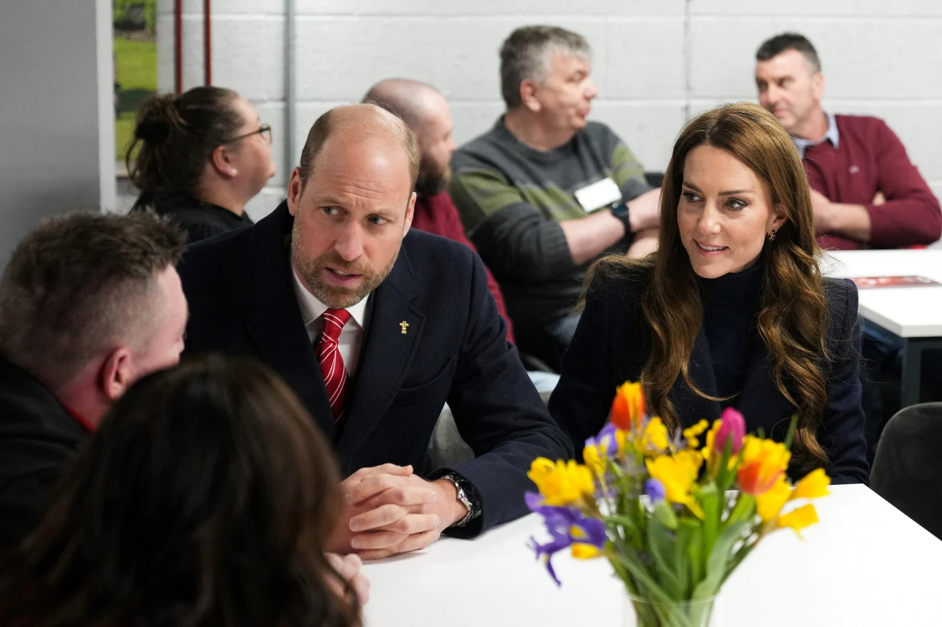 Para w eleganckim stroju siedzi przy stole w otoczeniu kilku innych osób, na środku stołu stoi wazon z kolorowymi kwiatami, w tle szare ściany i rozmawiające osoby.