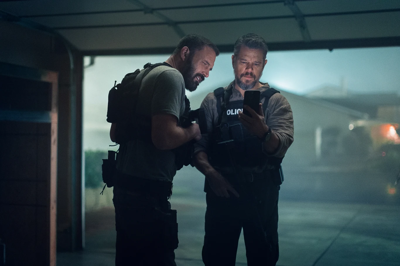 Dwóch umundurowanych policjantów w taktycznych kamizelkach ogląda coś na ekranie telefonu, stojąc w półotwartym garażu, w tle widoczny jest zamglony fragment podjazdu i budynku.