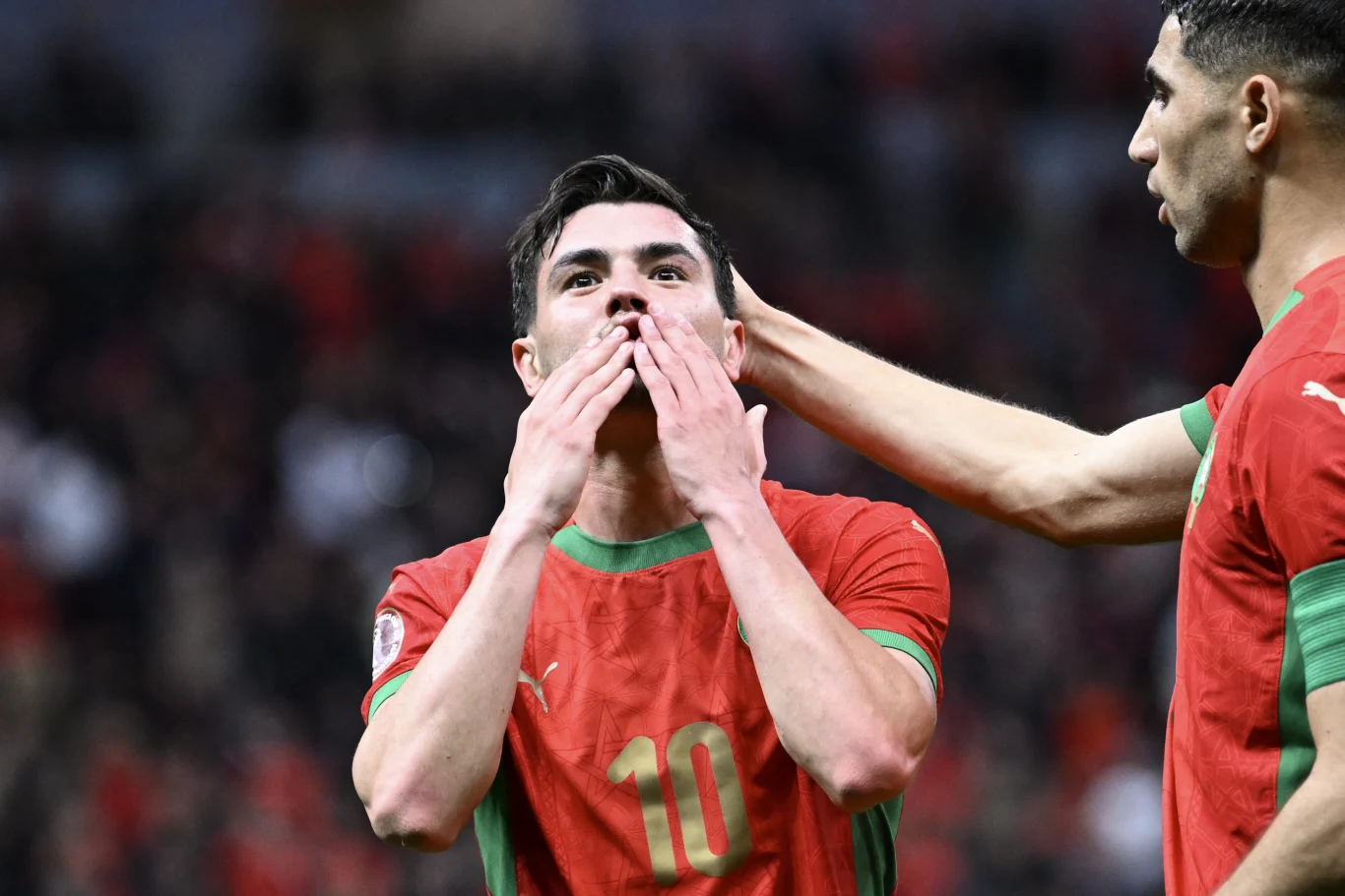 Piłkarz w czerwonej koszulce z numerem 10 przesyła pocałunki w stronę widowni, a jego kolega z drużyny dotyka go w geście wsparcia. Obaj mają na sobie stroje sportowe, w tle widoczna rozmyta publiczność stadionowa.