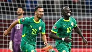 Dwóch piłkarzy reprezentacji Senegalu w zielonych strojach cieszy się z sukcesu na boisku, w tle widoczny bramkarz stojący przy bramce.