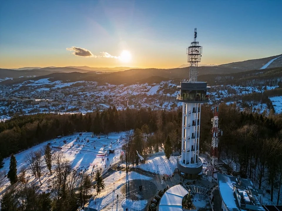 Wieża widokowa i przekaźnikowa otoczona zimowym lasem, śnieżna polana z ludźmi korzystającymi z atrakcji, w tle panorama miasta oraz gór oświetlonych zachodzącym słońcem.