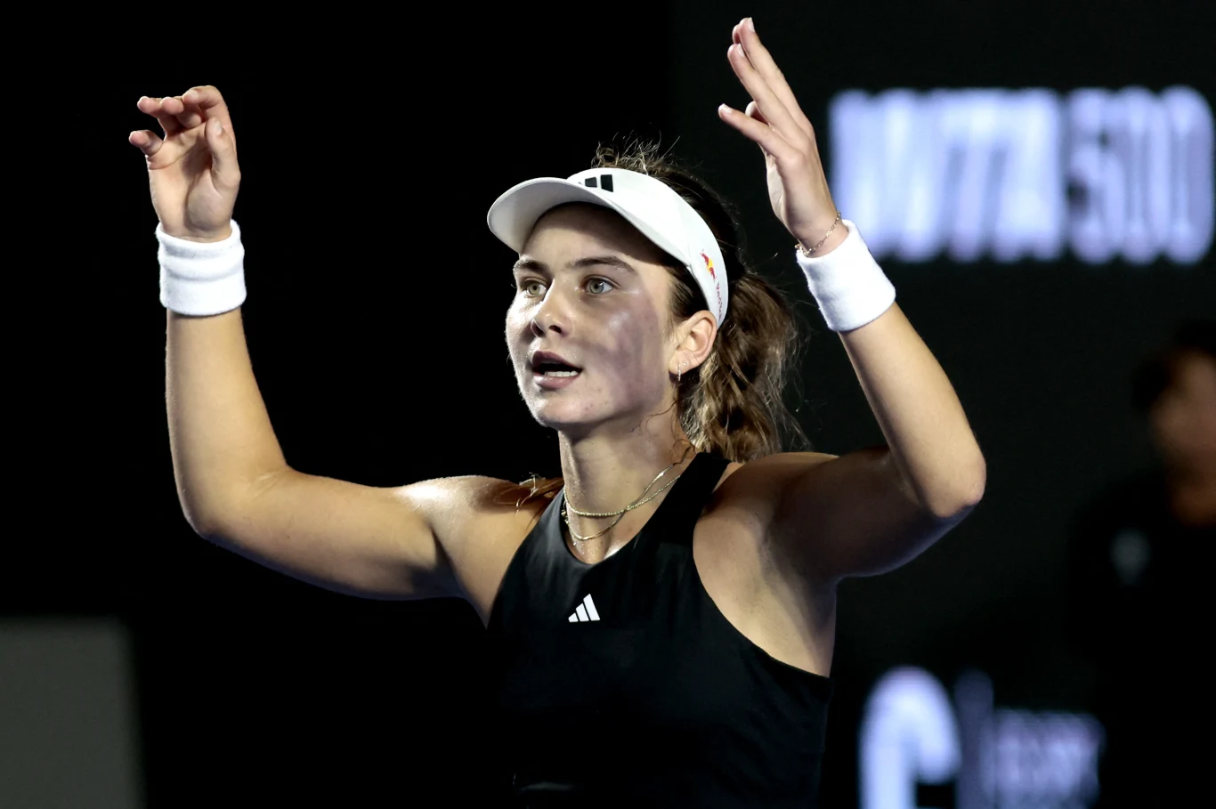 Młoda kobieta w sportowym stroju i białej czapce z daszkiem unosi ręce, wygląda na skoncentrowaną; na tle widoczne rozmyte logo turnieju tenisowego.