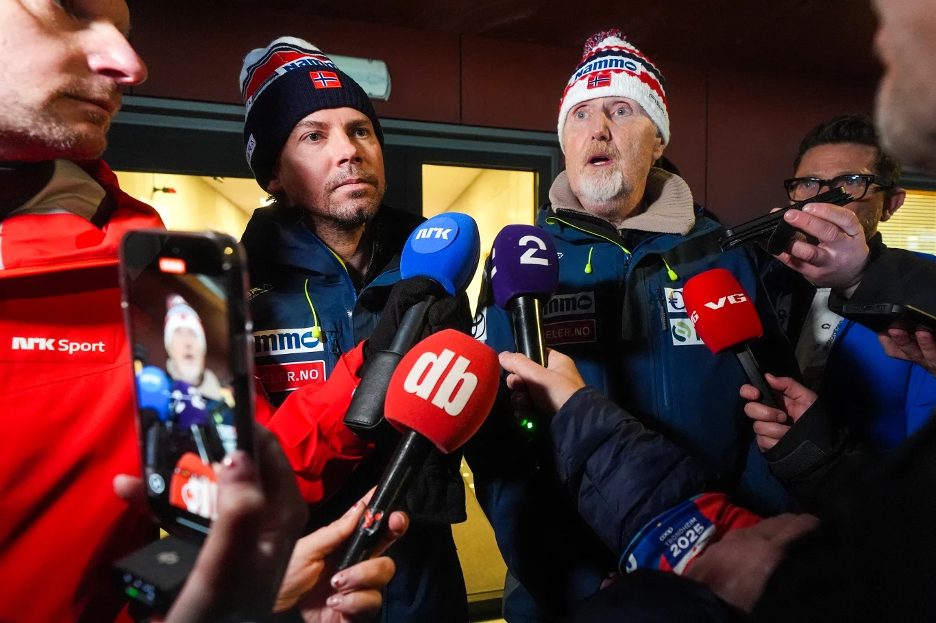 Dwóch mężczyzn w zimowych kurtkach i czapkach z logo norweskiej drużyny udziela wywiadu otoczonych przez dziennikarzy i mikrofony wielu stacji telewizyjnych. Na pierwszym planie widoczne są mikrofony z logotypami stacji i telefony nagrywające rozmowę.