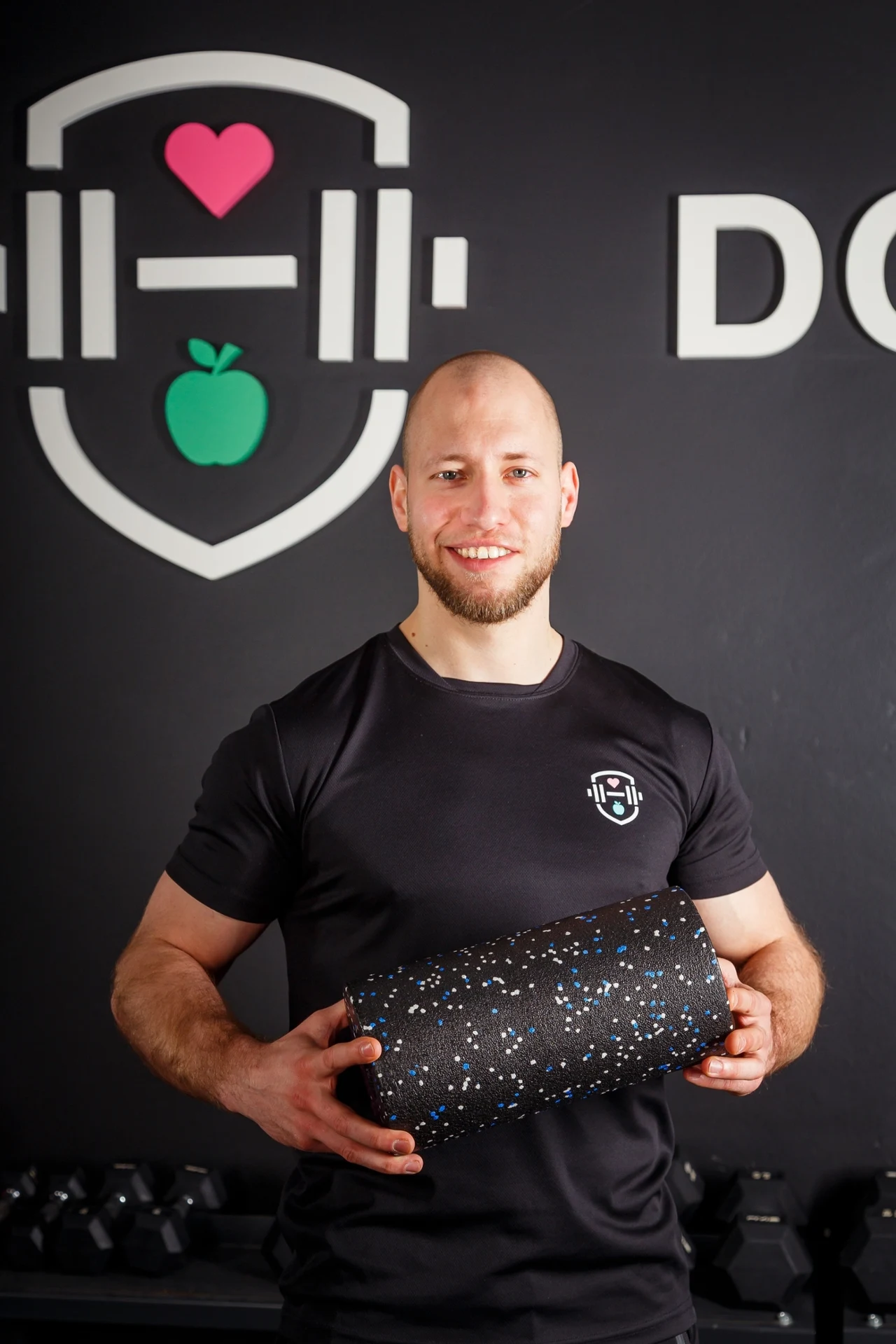 Uśmiechnięty mężczyzna w sportowej koszulce trzyma wałek do masażu, stojąc na tle czarnej ściany z dużym sportowym logotypem z różowym sercem i zielonym jabłkiem oraz hantlami ustawionymi w tle.