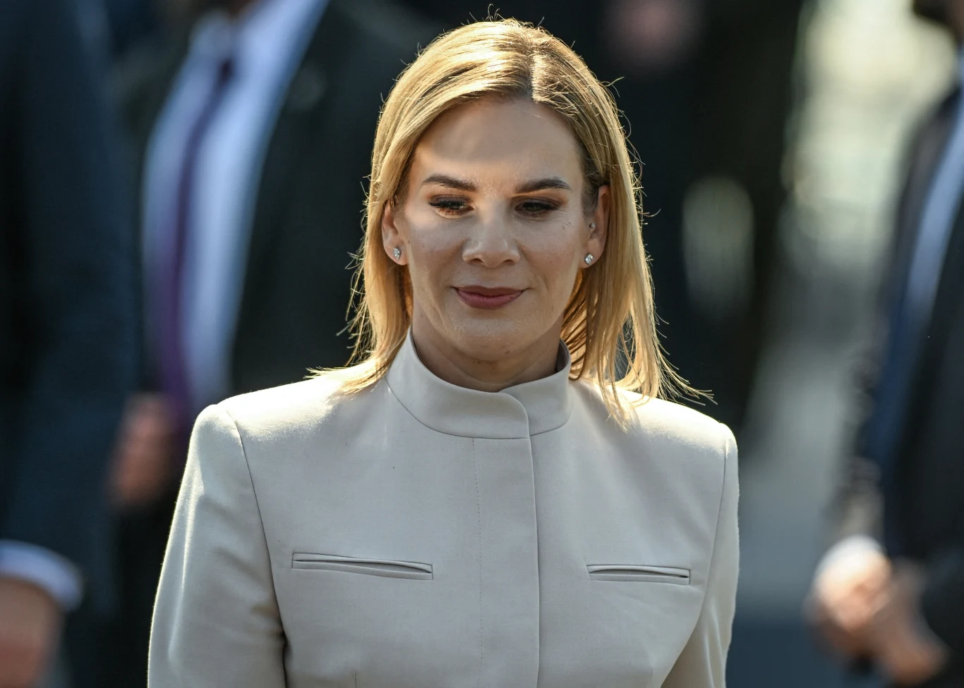 Kobieta o blond włosach w jasnej marynarce uśmiecha się subtelnie w kierunku aparatu. W tle widoczne są rozmyte sylwetki innych osób, skupienie pozostaje na głównej bohaterce zdjęcia.