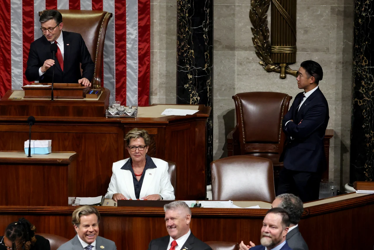 Wnętrze sali parlamentarnej, kilka osób w formalnych strojach prowadzi obrady, jedna osoba stoi przy fotelu przewodniczącego, duża flaga Stanów Zjednoczonych w tle, reszta uczestników siedzi i rozmawia.