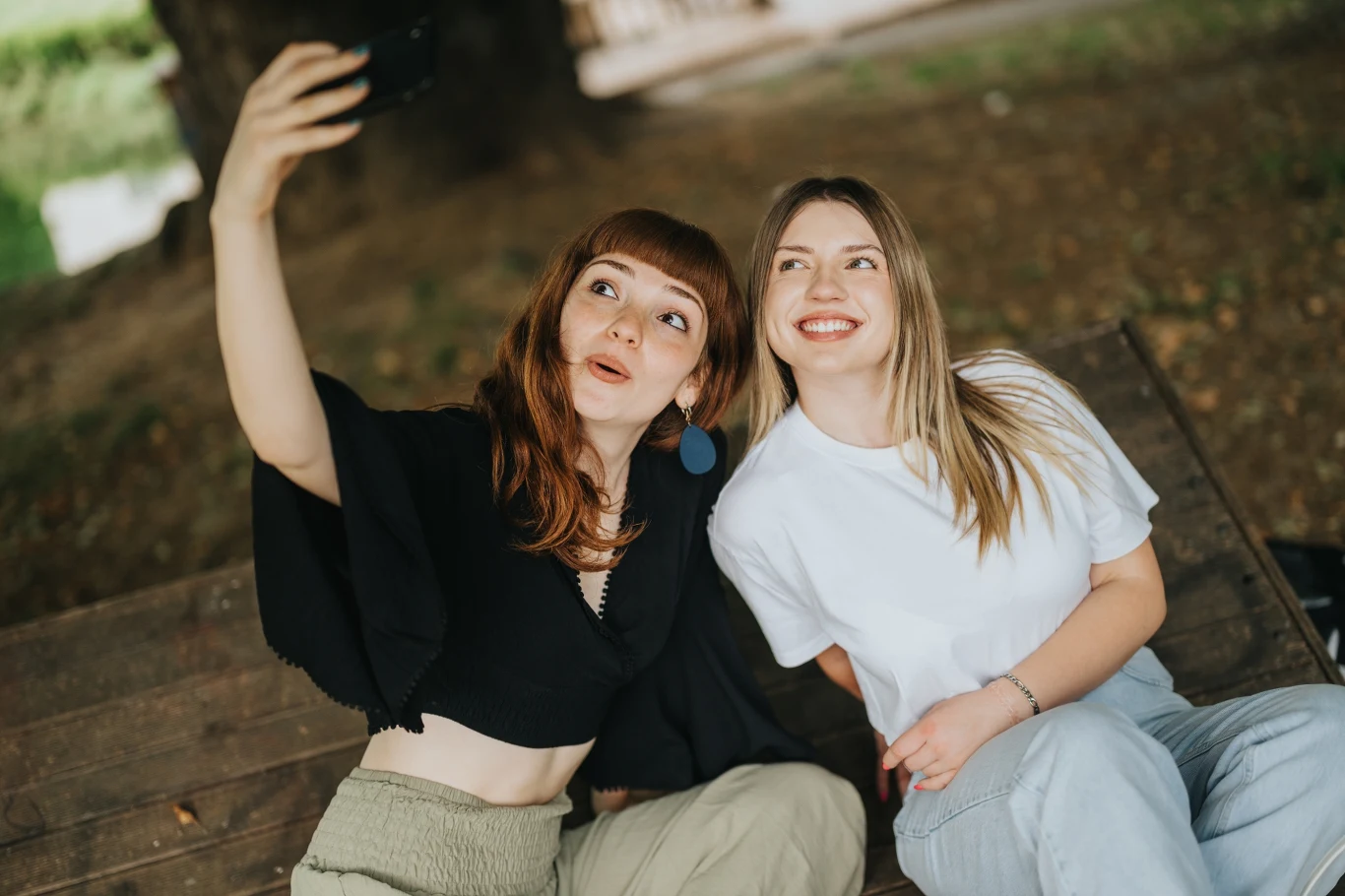 Dwie młode kobiety siedzą na drewnianej ławce w parku, robią sobie wspólne selfie. Jedna z nich uśmiecha się szeroko, a druga gestykuluje dłonią, trzymając telefon. W tle rozmyte drzewa i naturalne otoczenie.