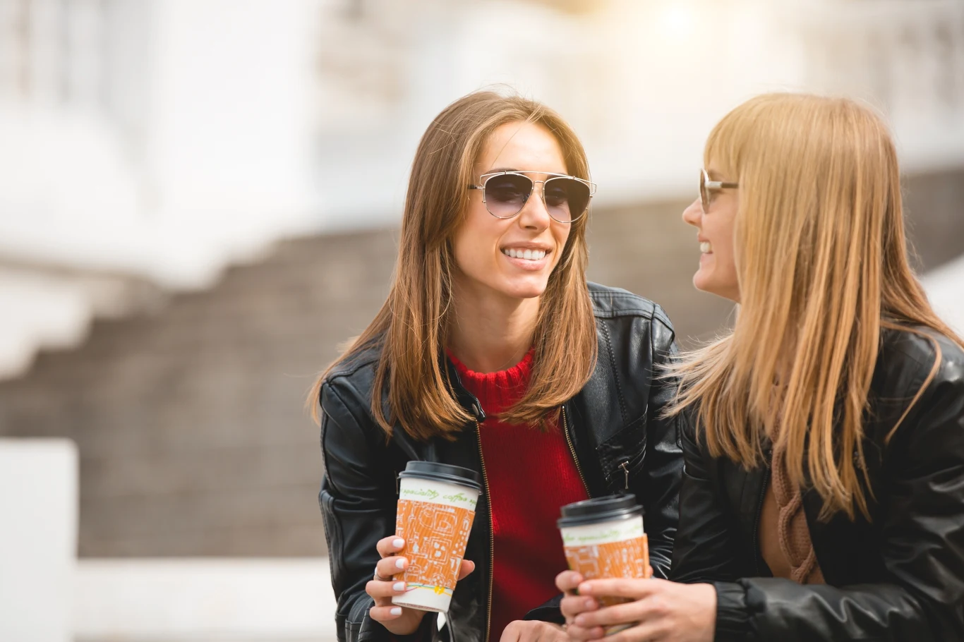 Dwie młode kobiety ubrane w ciemne kurtki i okulary przeciwsłoneczne rozmawiają, trzymając kubki z kawą na zewnątrz w pogodny dzień, uśmiechnięte i zrelaksowane