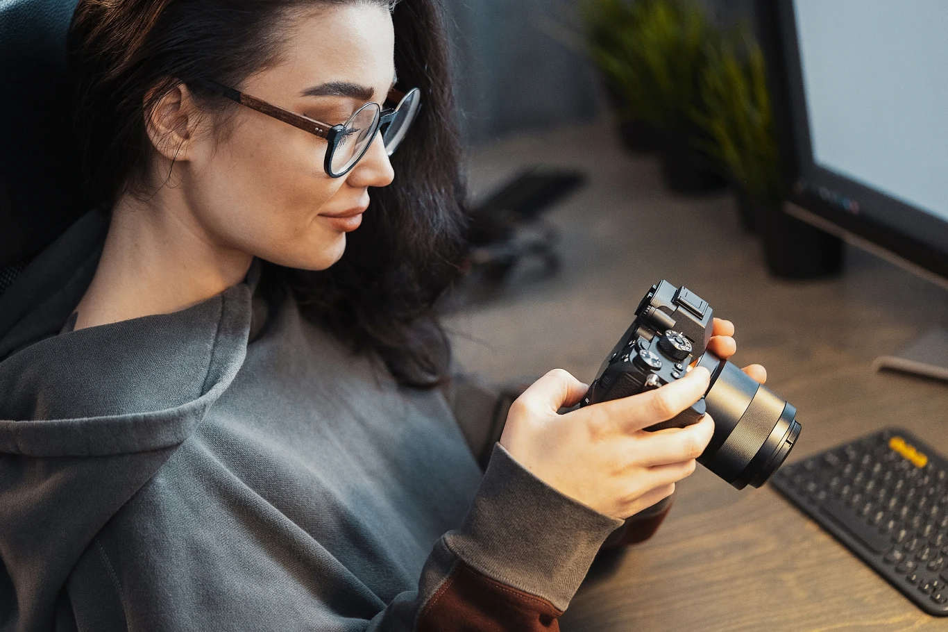 Kobieta z długimi ciemnymi włosami i okularami trzyma aparat fotograficzny w dłoniach w pobliżu biurka z klawiaturą komputerową i monitorem, skupiona na przeglądaniu zdjęcia lub ustawienia aparatu.