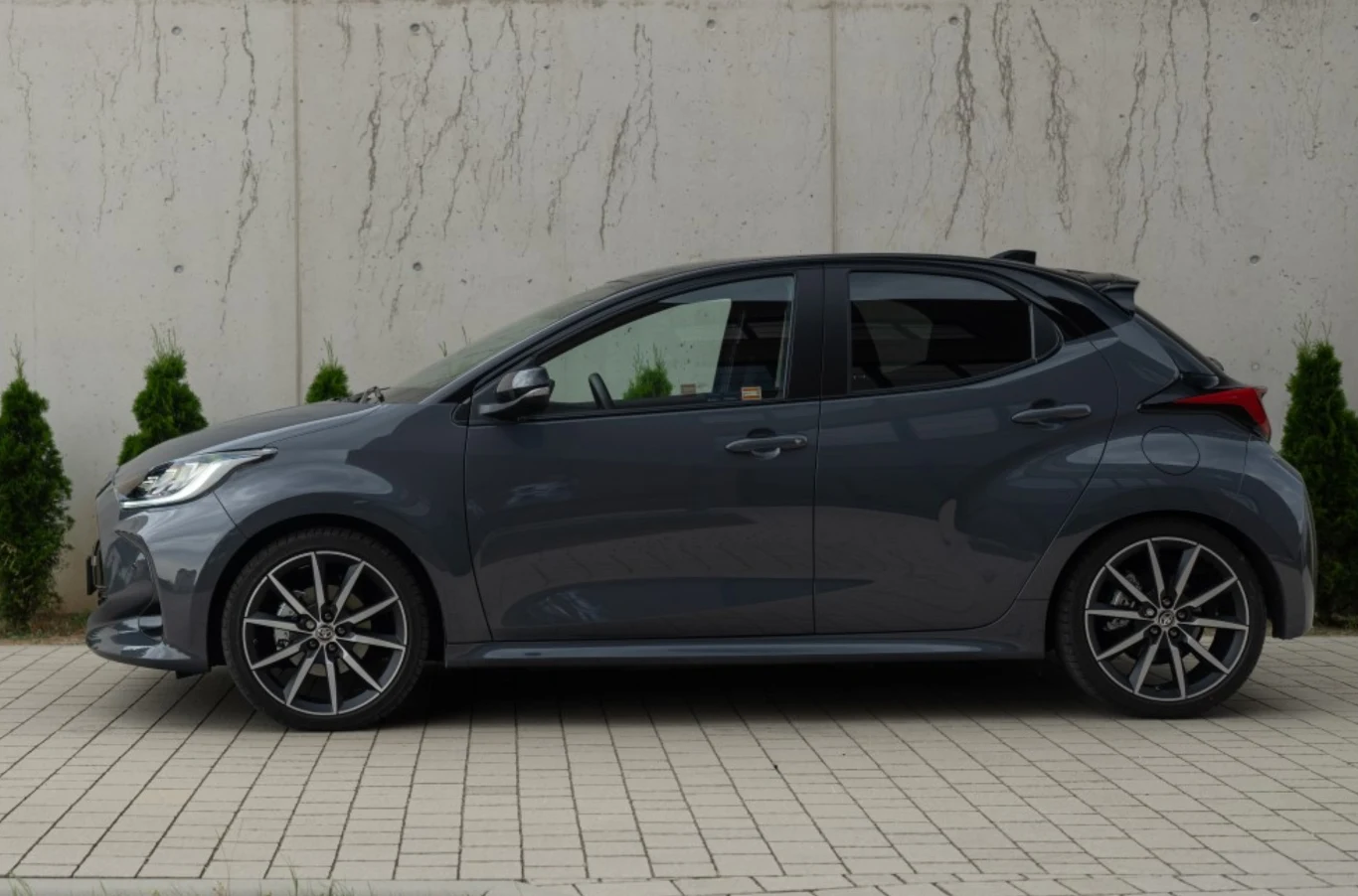 Szary hatchback zaparkowany na kostce brukowej przy ścianie z betonowych płyt, w tle zielone tuje równo rozmieszczone przy murze.