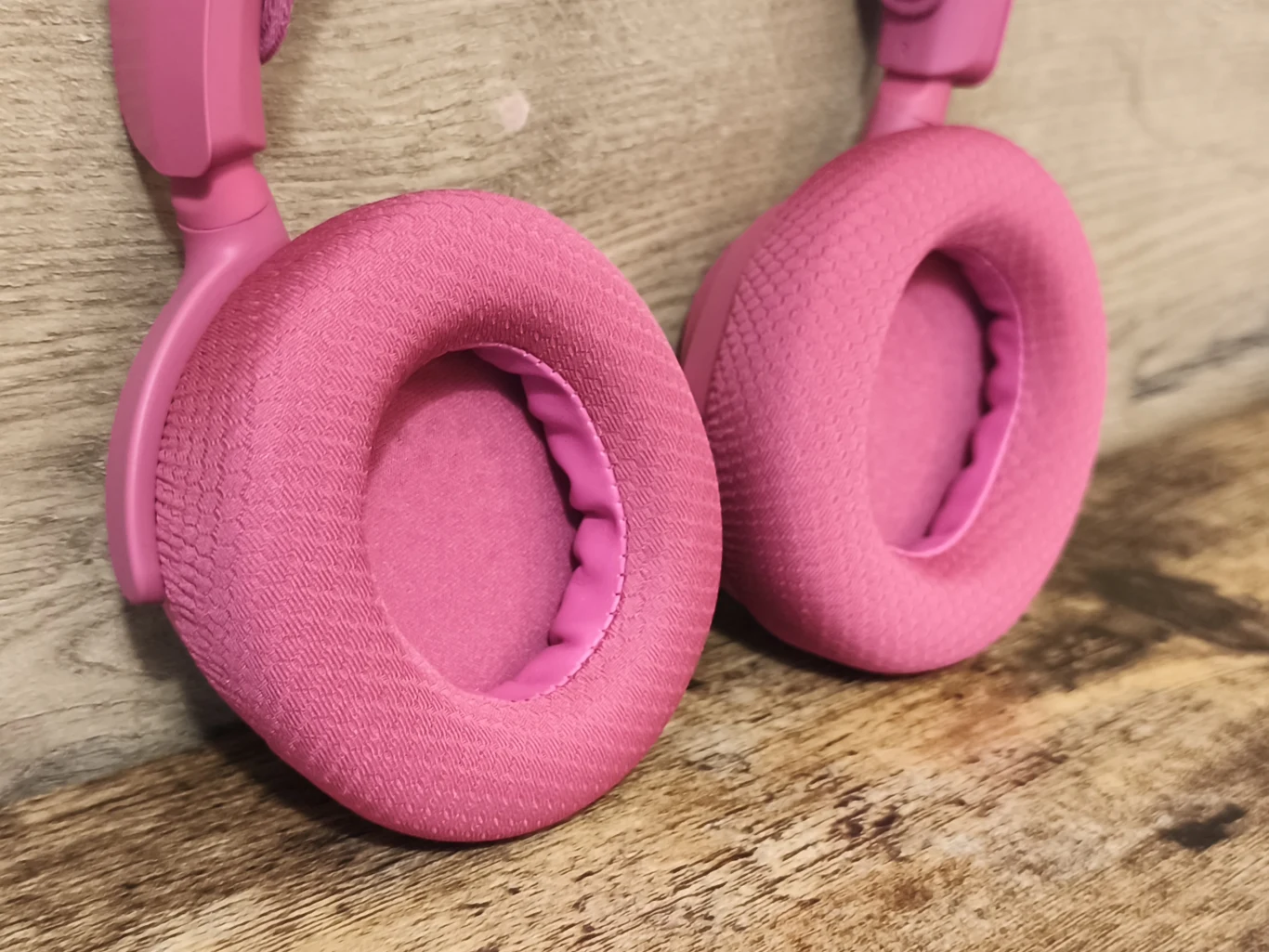 Różowe słuchawki z miękkimi, materiałowymi nausznicami ustawione na drewnianej powierzchni, widoczna faktura materiału oraz detal wykonania słuchawek.