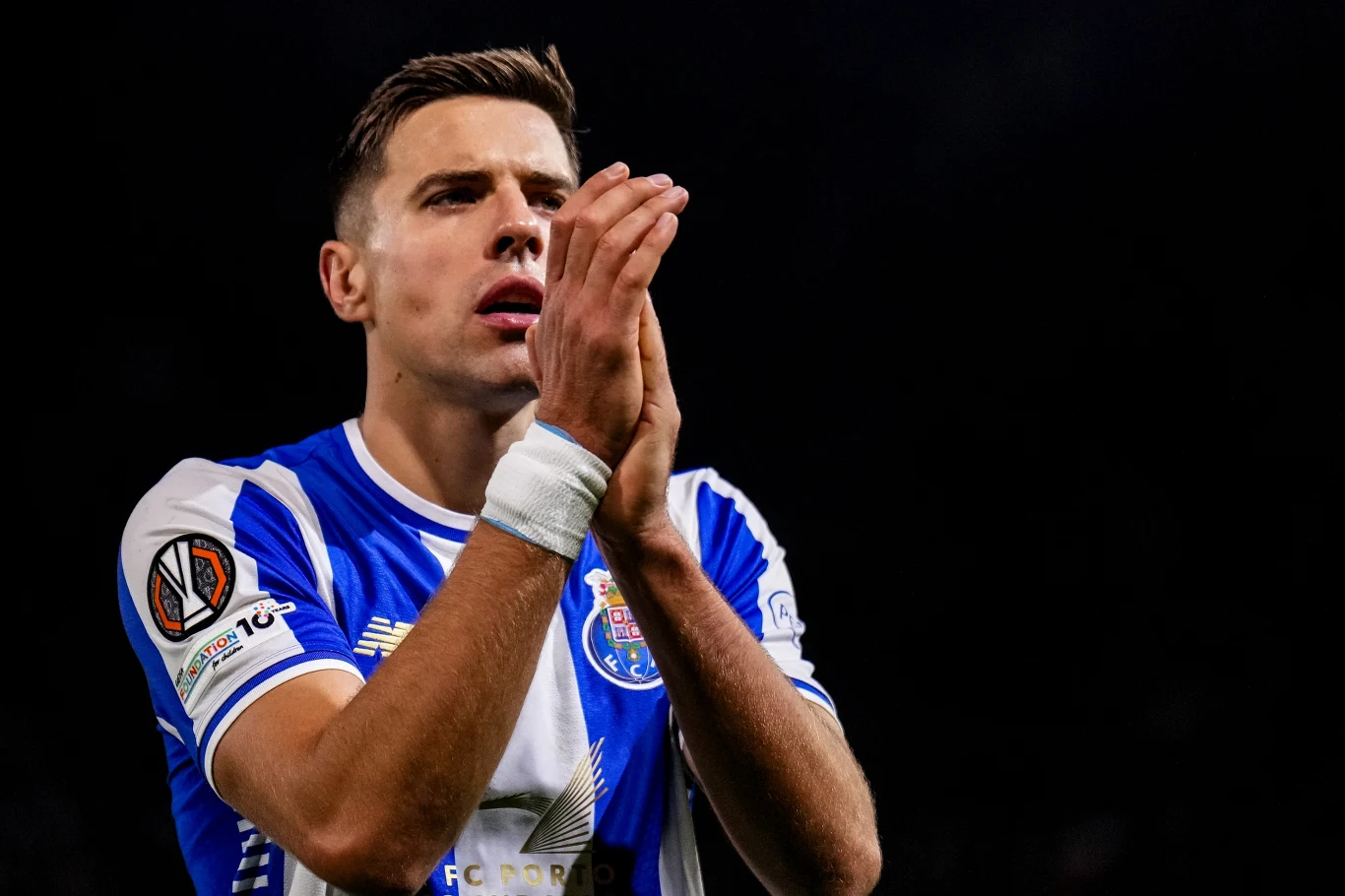 Jan Bednarek Piłkarz w niebiesko-białej koszulce z emblematami klubowymi i logiem Ligi Europy stoi ze złożonymi dłońmi, prawdopodobnie wyrażając emocje po meczu na stadionie, tło jest ciemne.