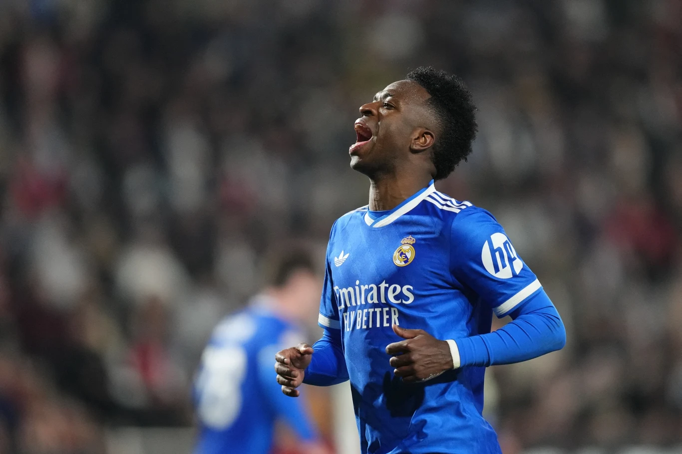 Piłkarz w niebieskim stroju Realu Madryt z wyraźnym wyrazem emocji podczas meczu na stadionie, w tle rozmyty tłum kibiców i inny zawodnik.