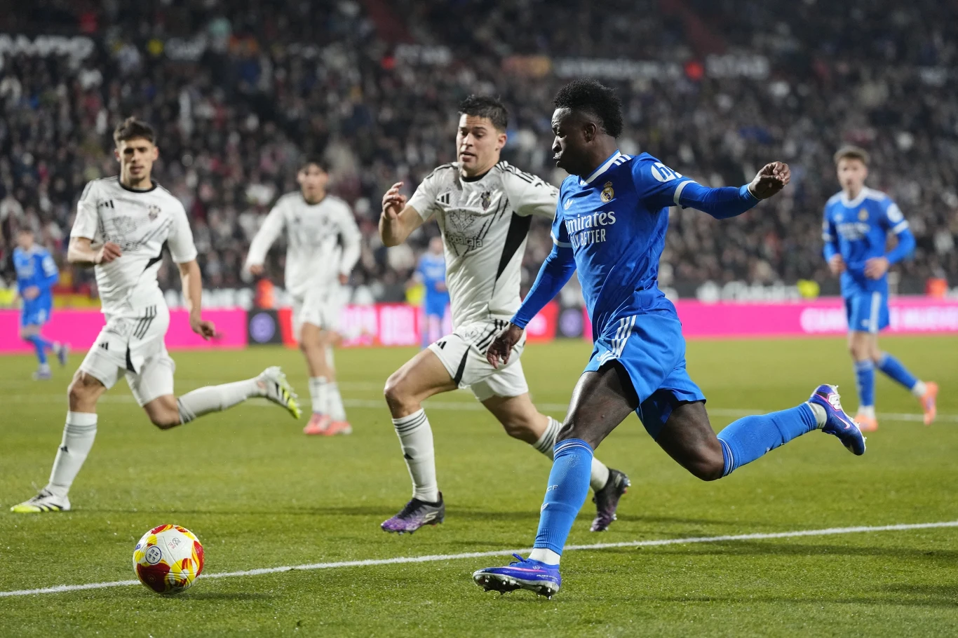 Zawodnik w niebieskim stroju oddaje strzał na bramkę podczas meczu piłkarskiego, wokół niego kilku piłkarzy drużyny w białych strojach oraz tłum kibiców na trybunach.