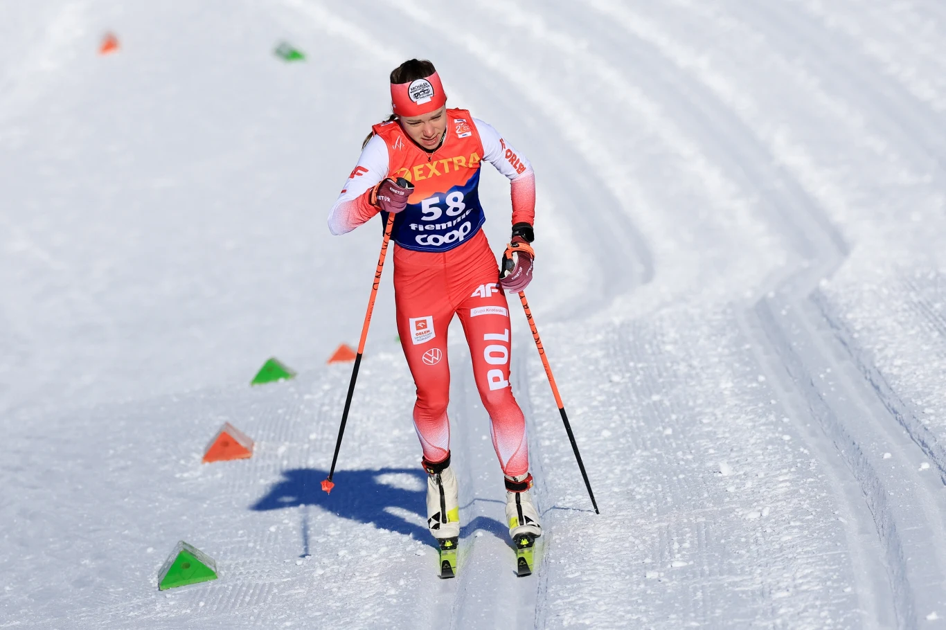 Biegaczka narciarska w czerwonym stroju reprezentacji Polski, w trakcie wyścigu na trasie narciarskiej, otoczona śniegiem oraz kolorowymi znacznikami na trasie.