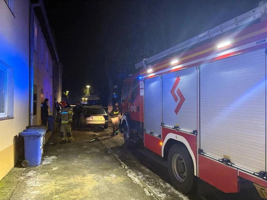 Tragedia w Chełmnie. Kobieta i jej dzieci nie żyją po zatruciu czadem Strażacy w mundurach oraz wóz strażacki stojący przy oświetlonej ulicy obok budynku mieszkalnego, obok zaparkowany samochód osobowy. W tle inne osoby i światła uliczne.