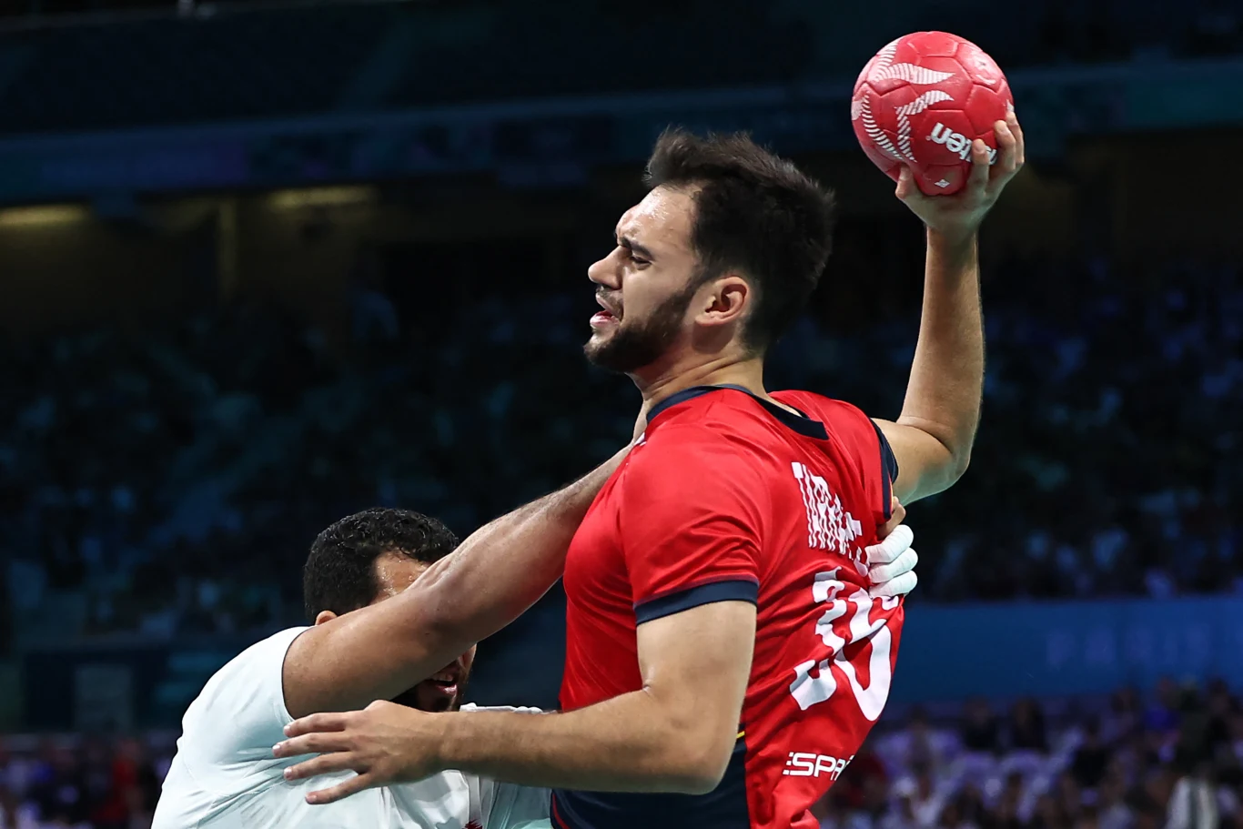 Zawodnik w czerwonym stroju z piłką ręczną uniesioną do rzutu, w tle przeciwnik próbujący go zatrzymać, dynamiczne ujęcie z widownią na trybunach.